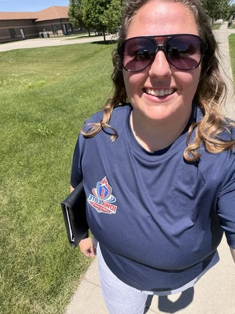 A smiling woman with sunglasses, wearing a blue sports shirt, standing on a grassy outdoor area with buildings and trees in the background.