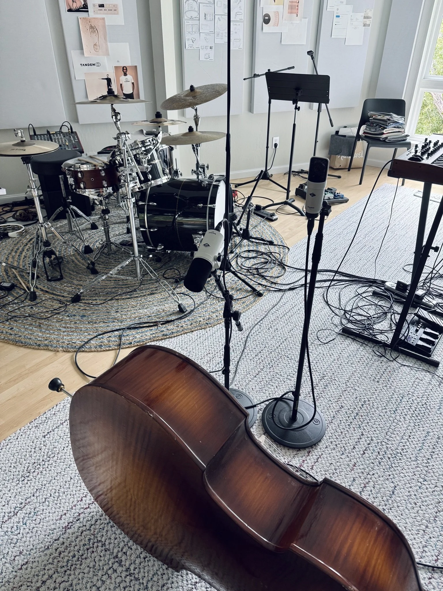 A music studio with a drum set, microphone, music stand, and a chair with stacks of papers, near a window with trees outside, and various posters and diagrams on the wall.