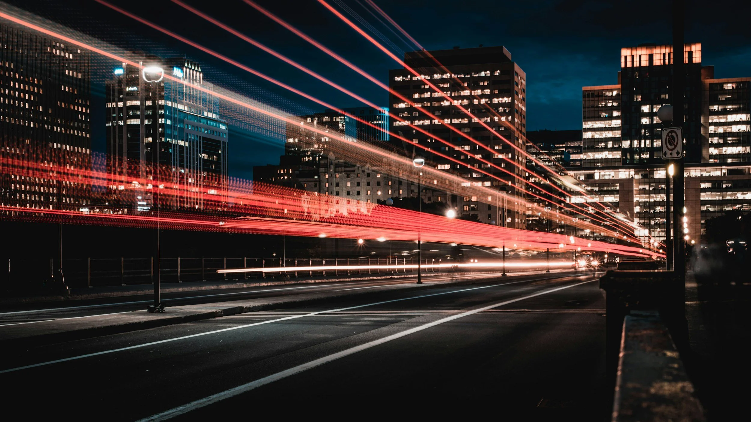 A cityscape at night with tall buildings illuminated, and light trails from moving vehicles on the road.