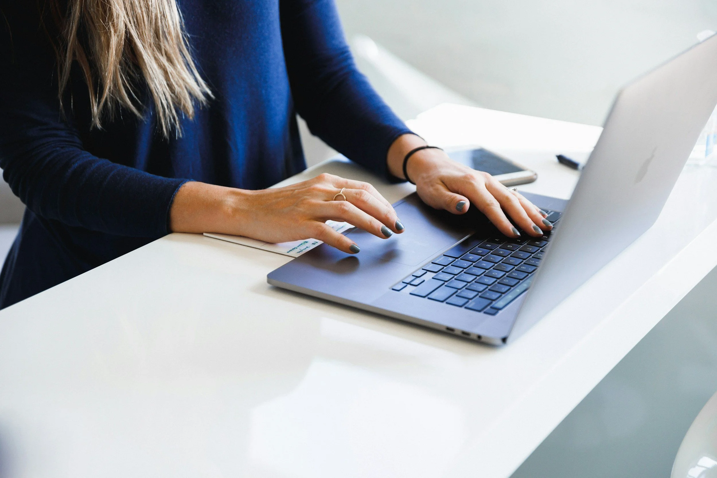 Close-up of a person typing on a laptop with a smartphone and paper on a white table.