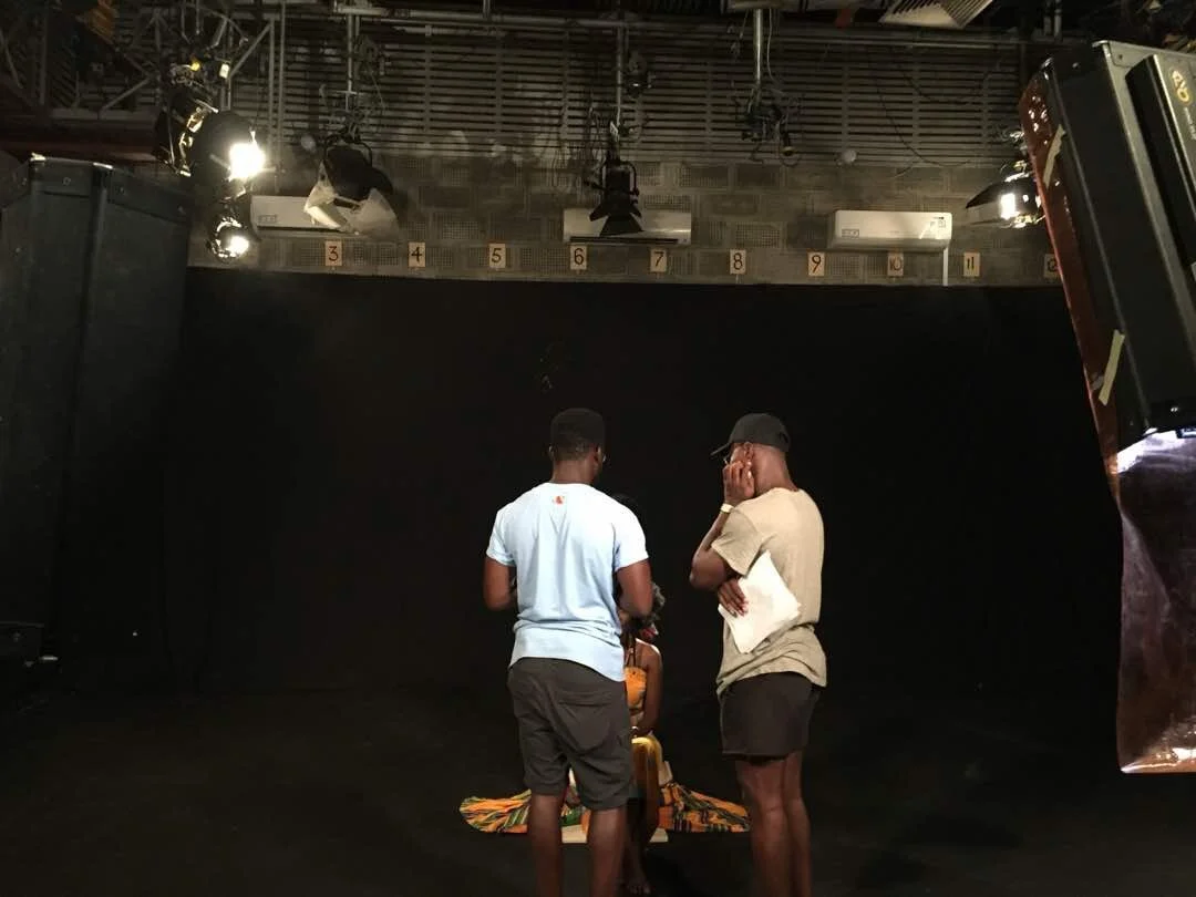 Three people in a studio with black walls and stage lights overhead. One person is sitting on a fabric, two others stand nearby engaging in conversation.