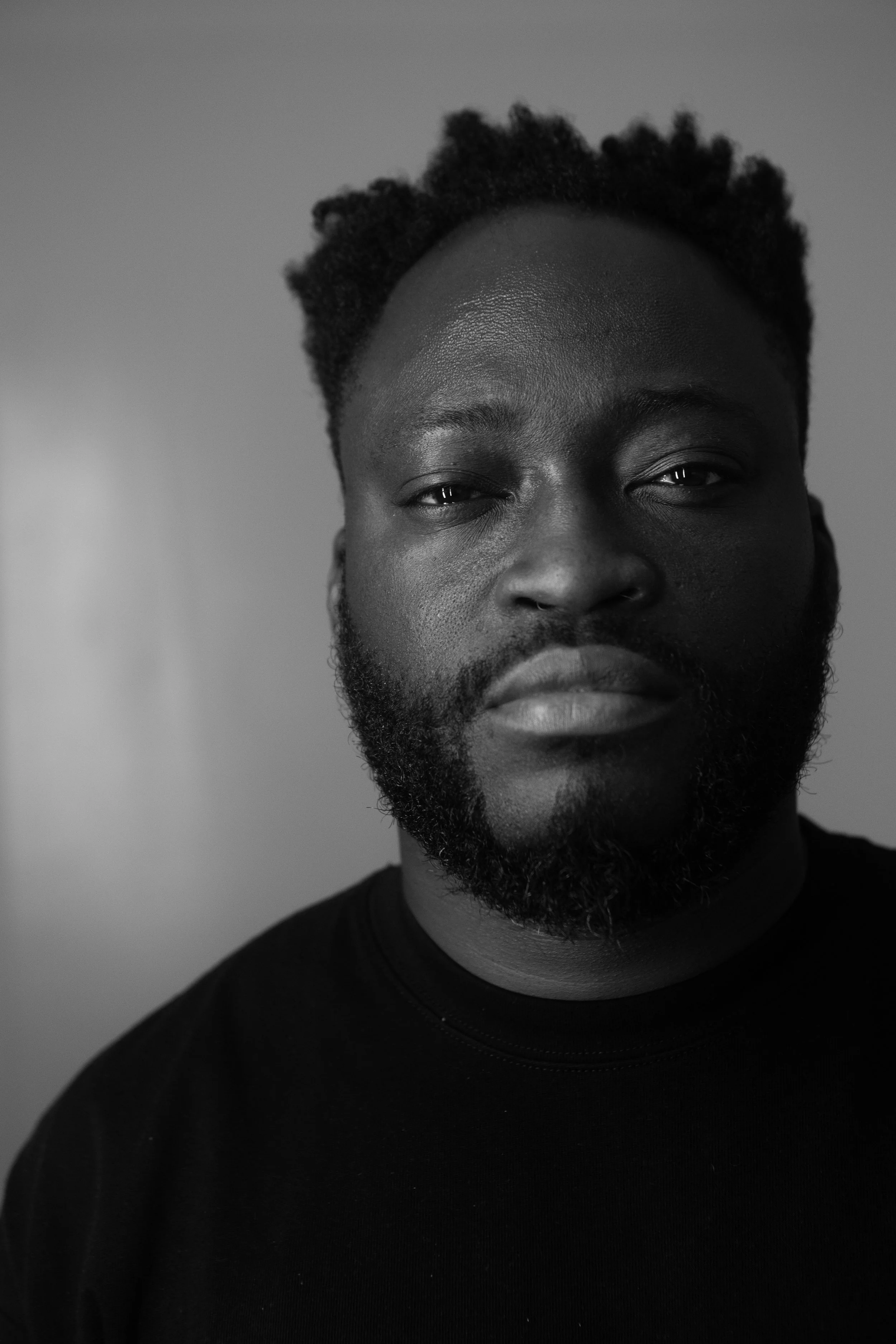 A black and white portrait photograph of a young man with dark skin, short curly hair, and a beard, wearing a black shirt and looking directly at the camera.
