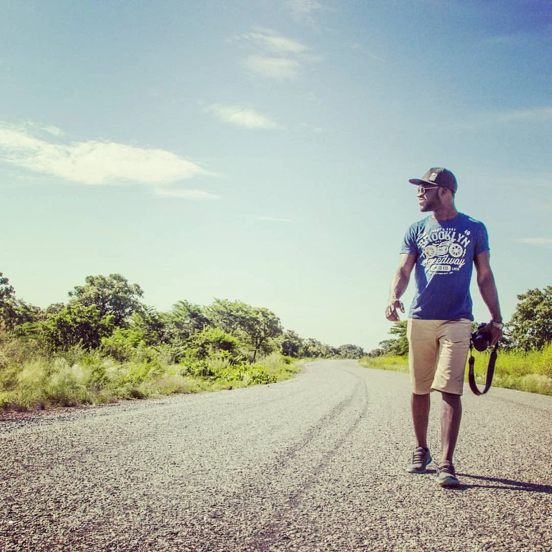 A man is walking on a gravel road in a rural area with green trees and bushes on both sides. He is wearing a blue t-shirt, beige shorts, a cap, sunglasses, and is holding a camera in his right hand. The sky is clear with a few clouds.