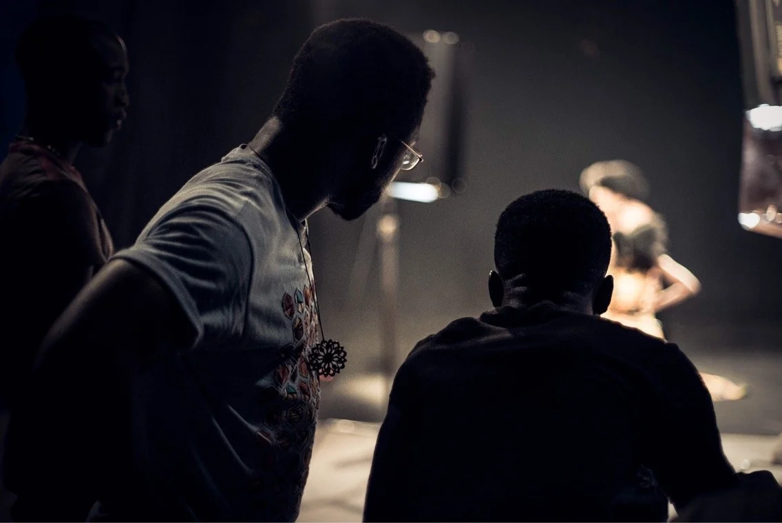 Two men in the foreground observing a scene on stage, with a person holding a microphone and a guitar in the background, during a rehearsal or performance.