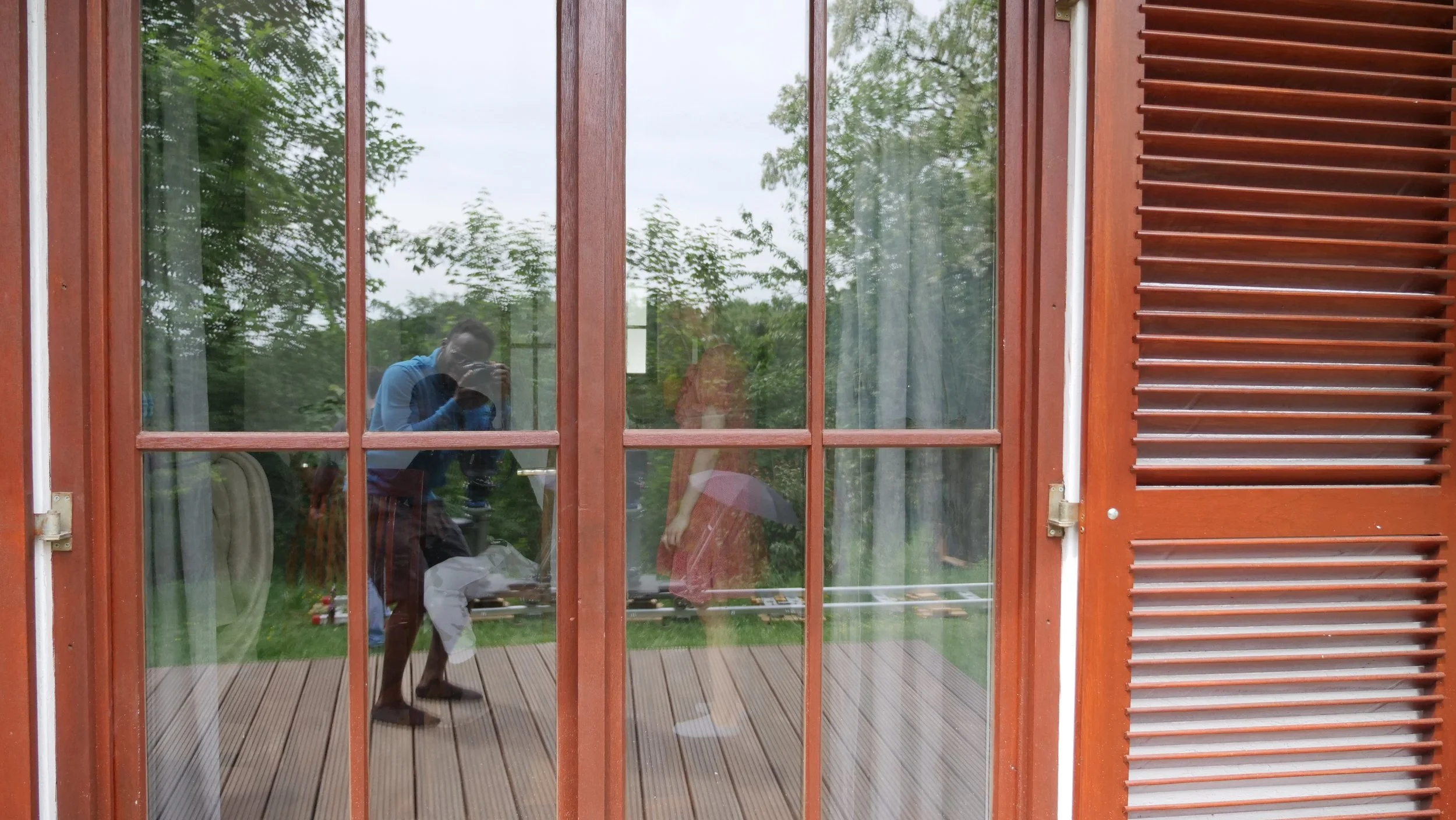 Reflection of two people taking a photo in a large window with brown wooden frames, overlooking a lush green yard.