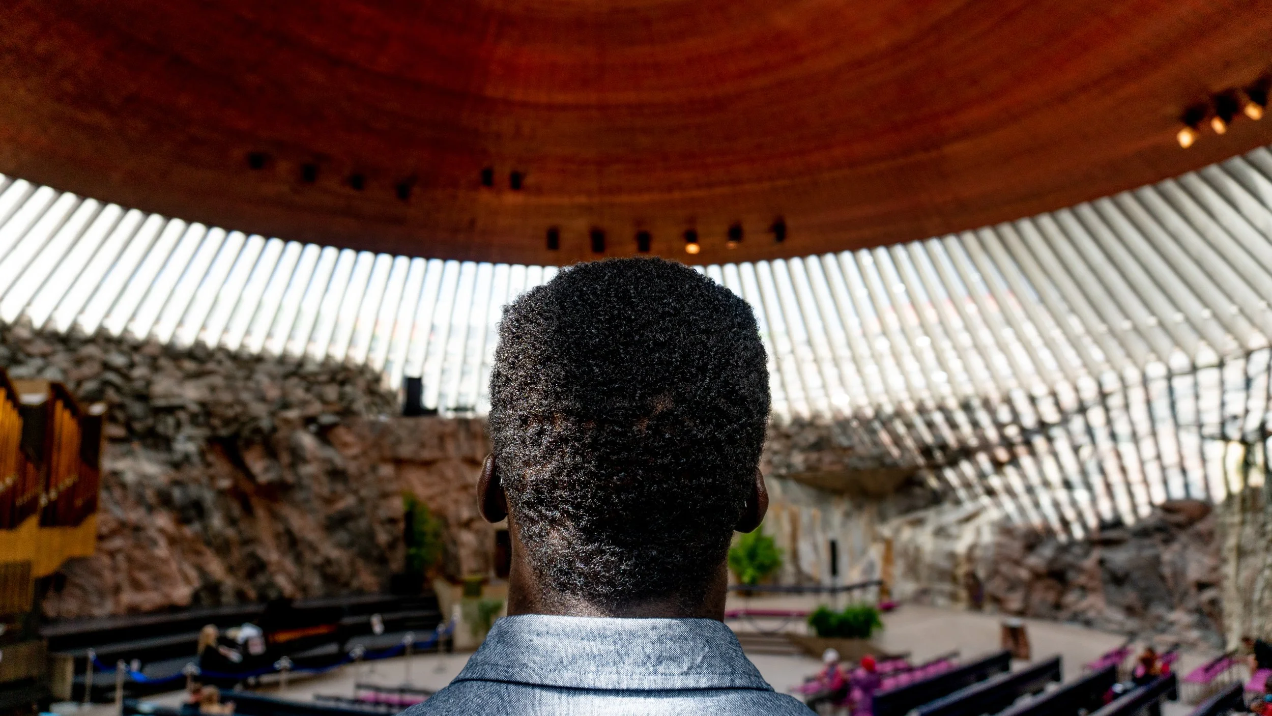 A person with short curly black hair seen from behind, standing inside a large, modern architectural structure with a wooden ceiling and tall, curved, white beams.