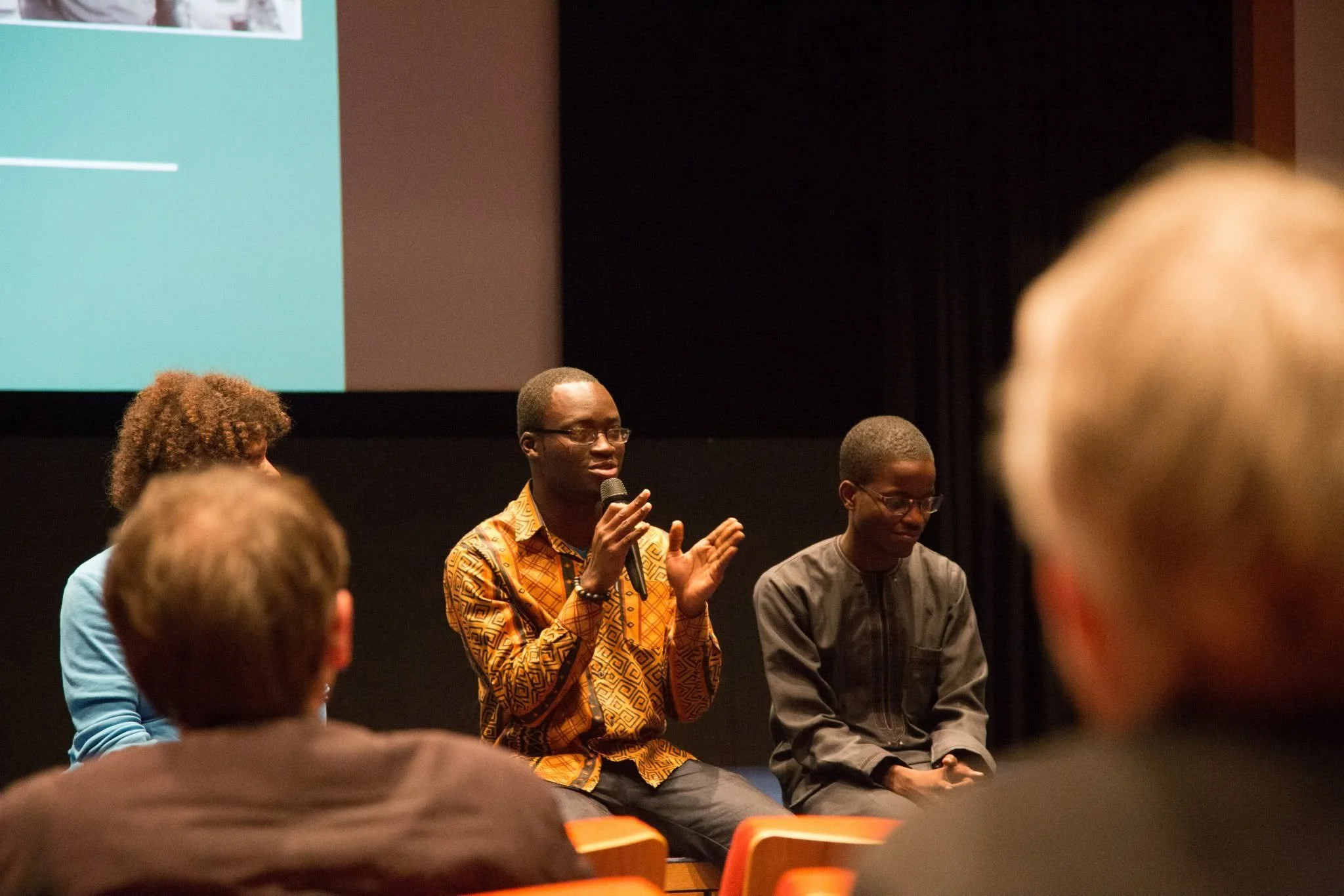 A man with glasses and patterned orange shirt speaking into a microphone, seated next to a person with glasses and a striped shirt, in front of an audience during a panel discussion or presentation.