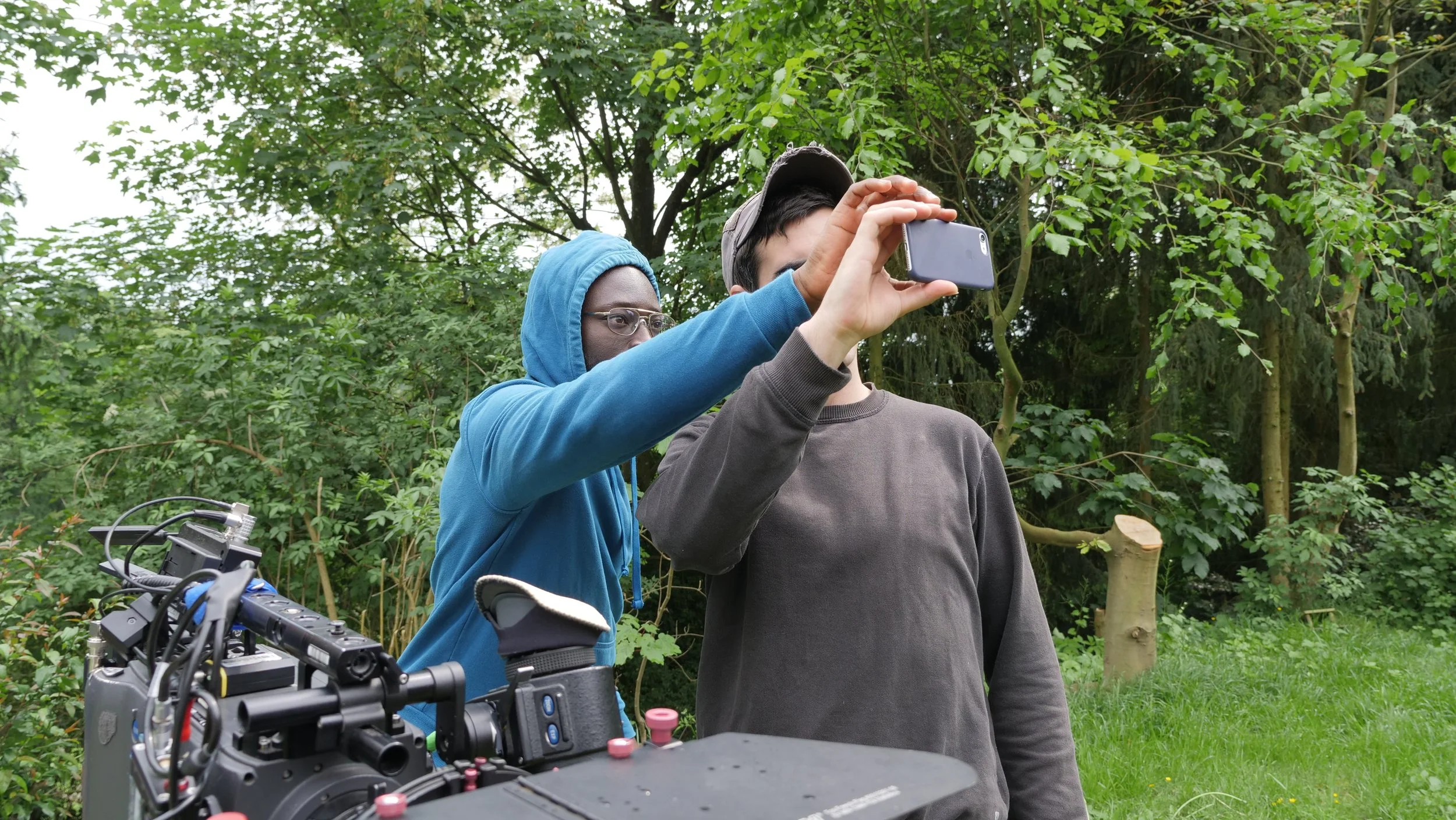 Two young men are outdoors among trees, one holding a smartphone and the other wearing a hoodie and glasses; there is filming equipment in the foreground.