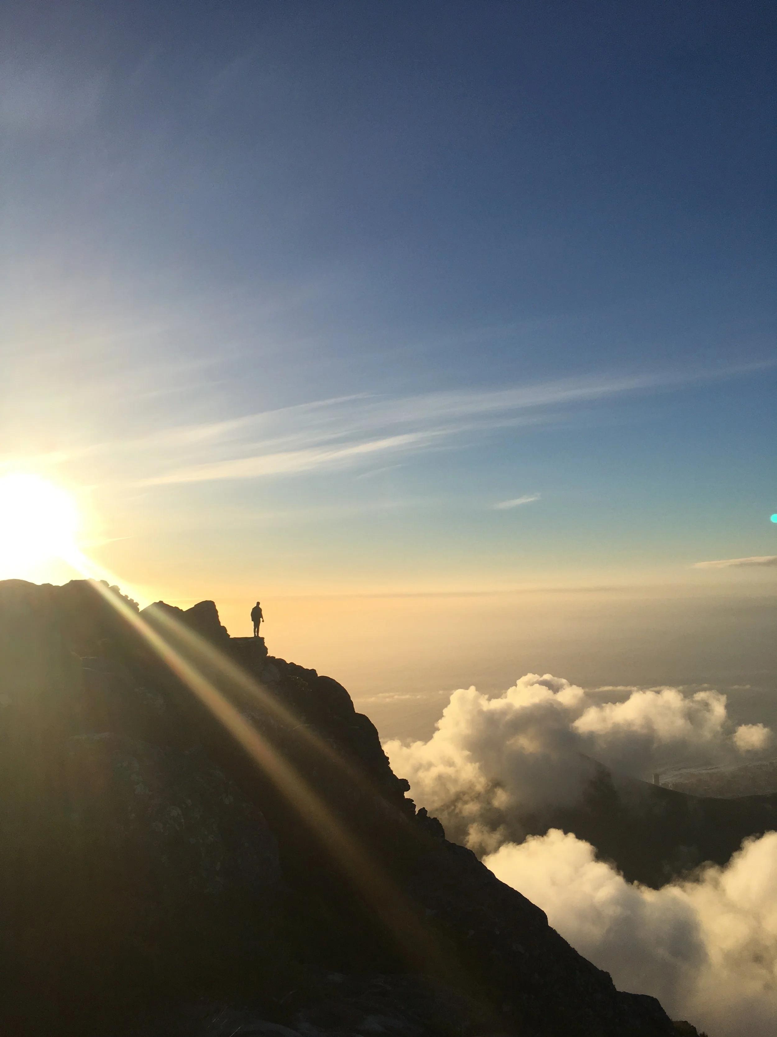 A person standing on a mountain ledge at sunset, with clouds below and a clear blue sky above.