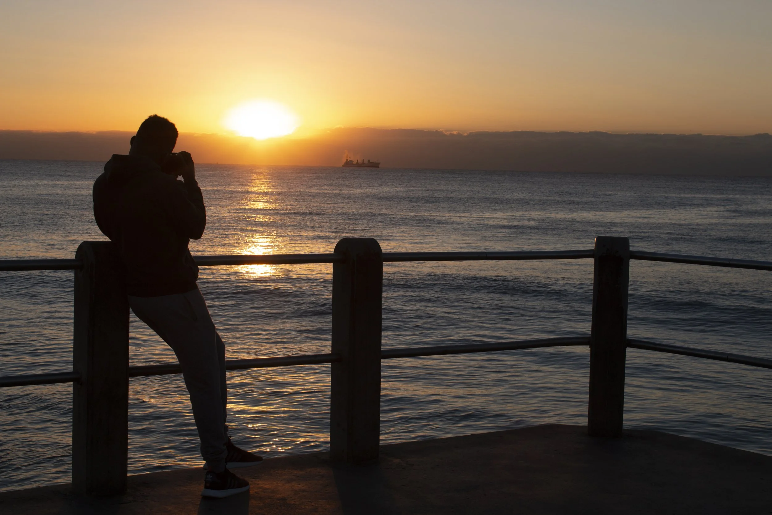 Silhouette of a person taking a photo of a sunset over the ocean with a cargo ship in the background.
