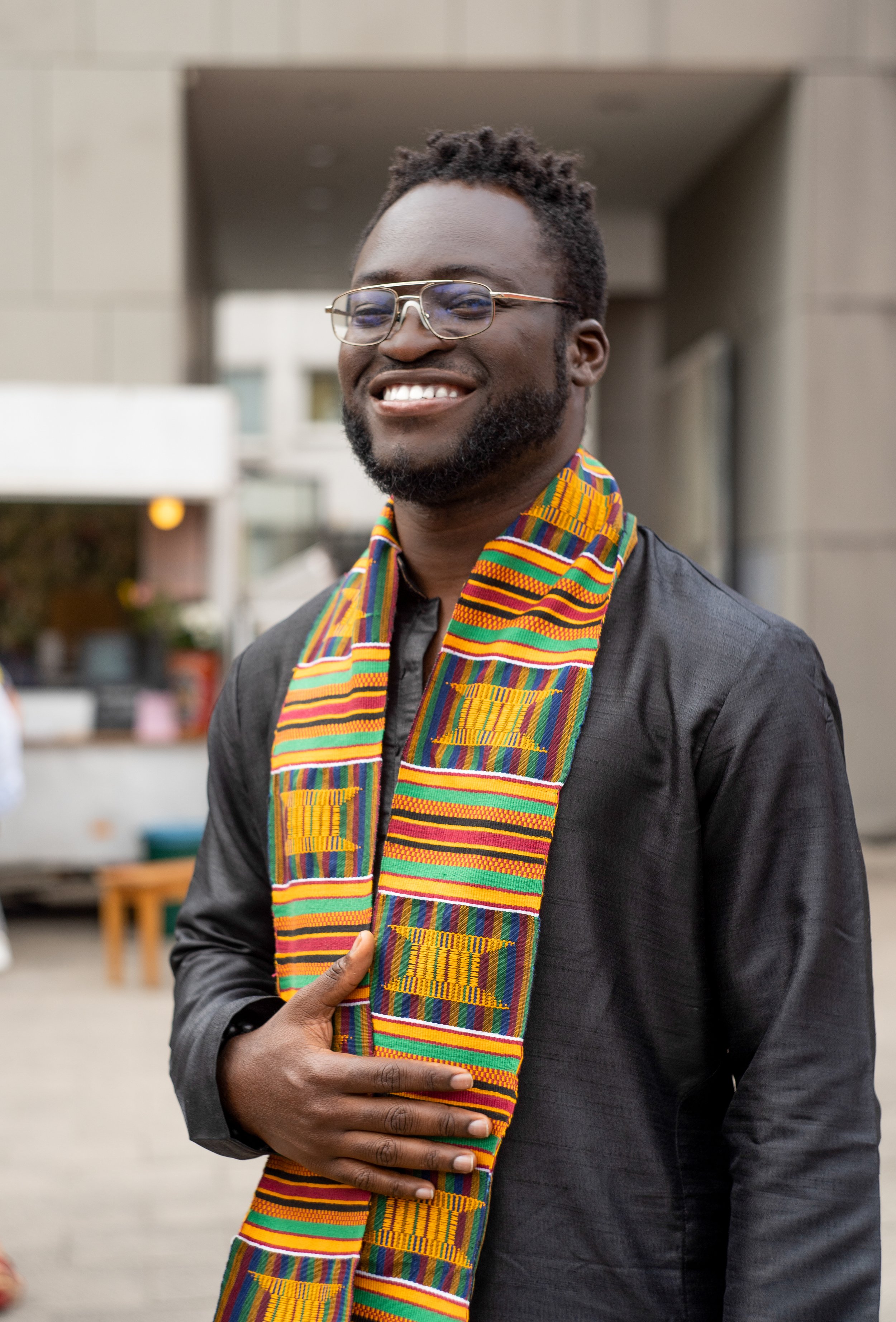 A smiling man with glasses and a beard wearing a colorful Kente cloth scarf, standing outdoors in front of a building.