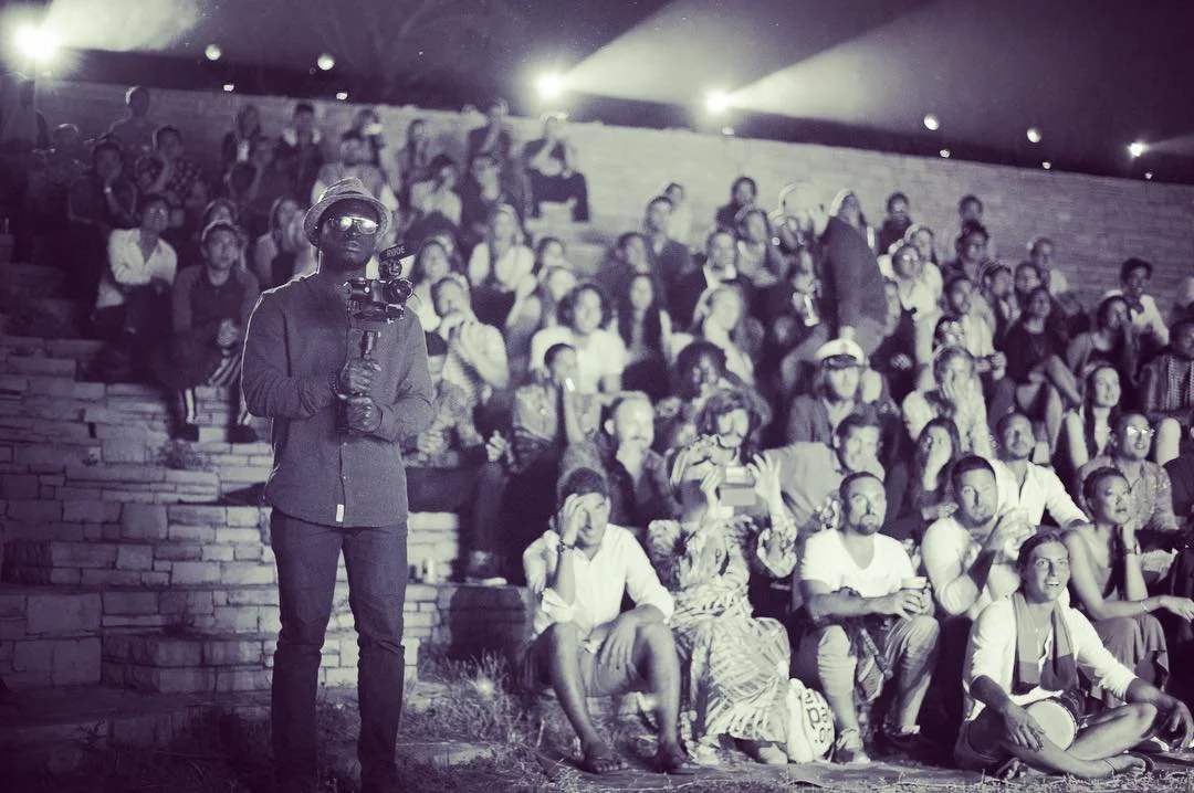 A man holding a video camera stands in front of an outdoor audience seated on tiered steps at night. The audience members are watching a performance or event, some clapping, taking photos, or smiling.