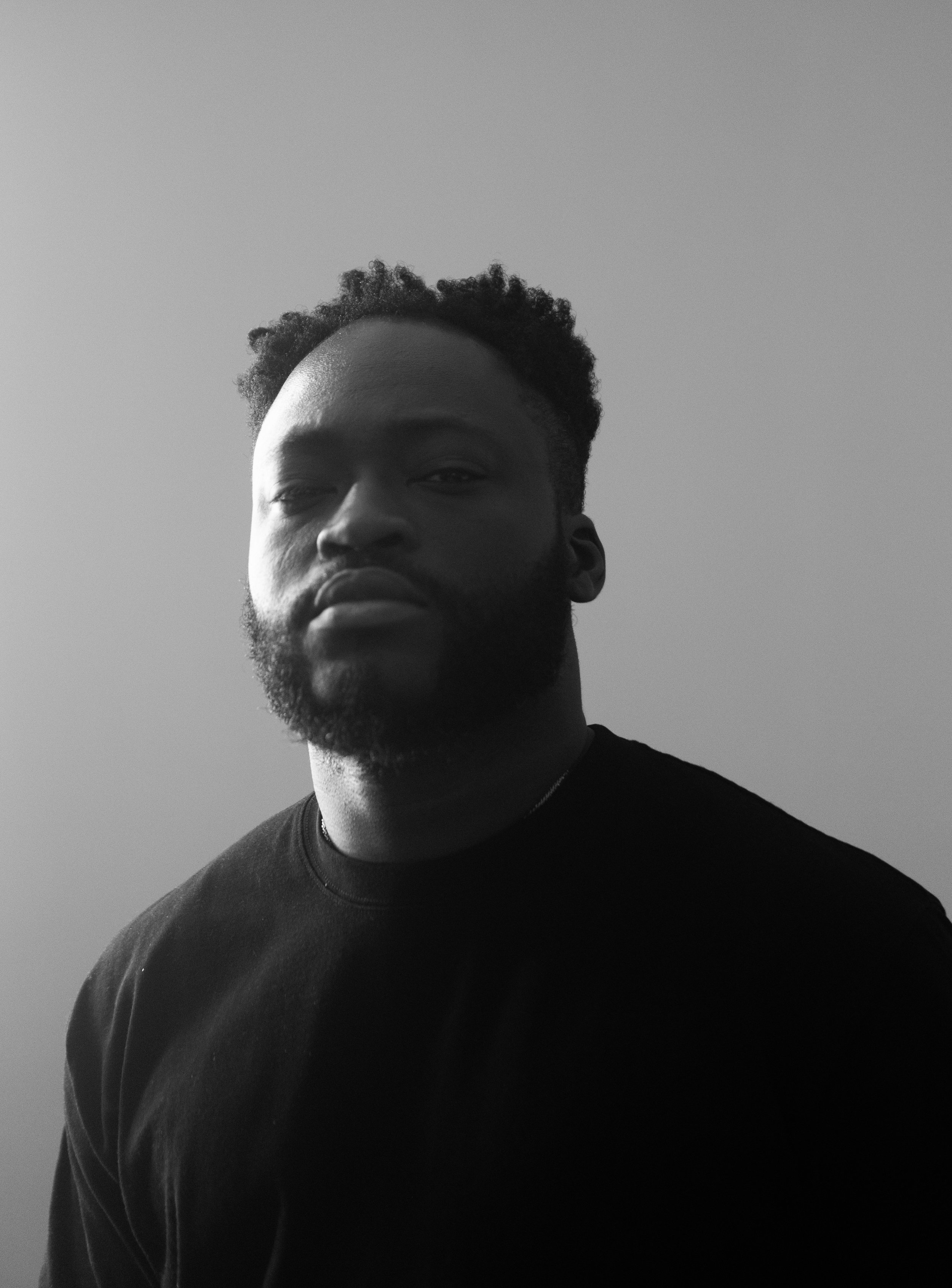 Black and white photograph of a young man with short curly hair and a beard, wearing a dark shirt, looking at the camera with a serious expression, against a plain gray background.