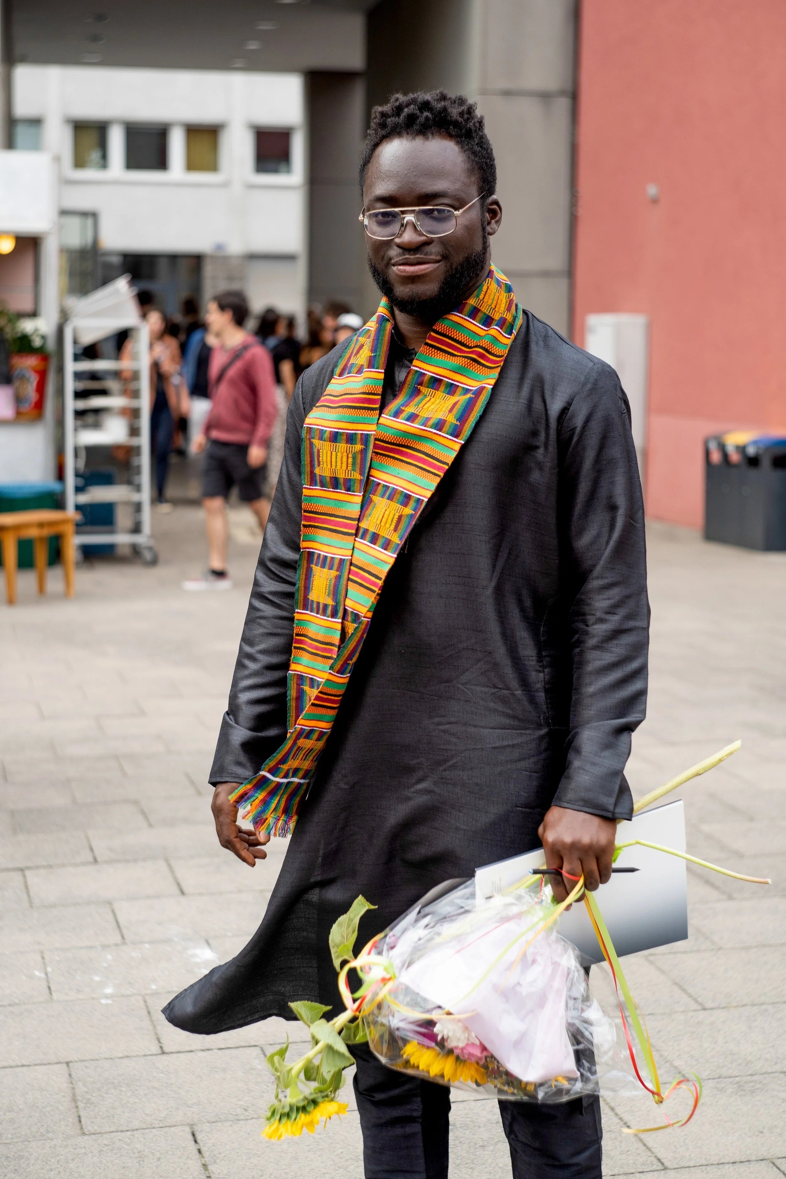 A man in black clothing and colorful African sash holding a bouquet of flowers and a gift bag, walking outside near a crowd and buildings.
