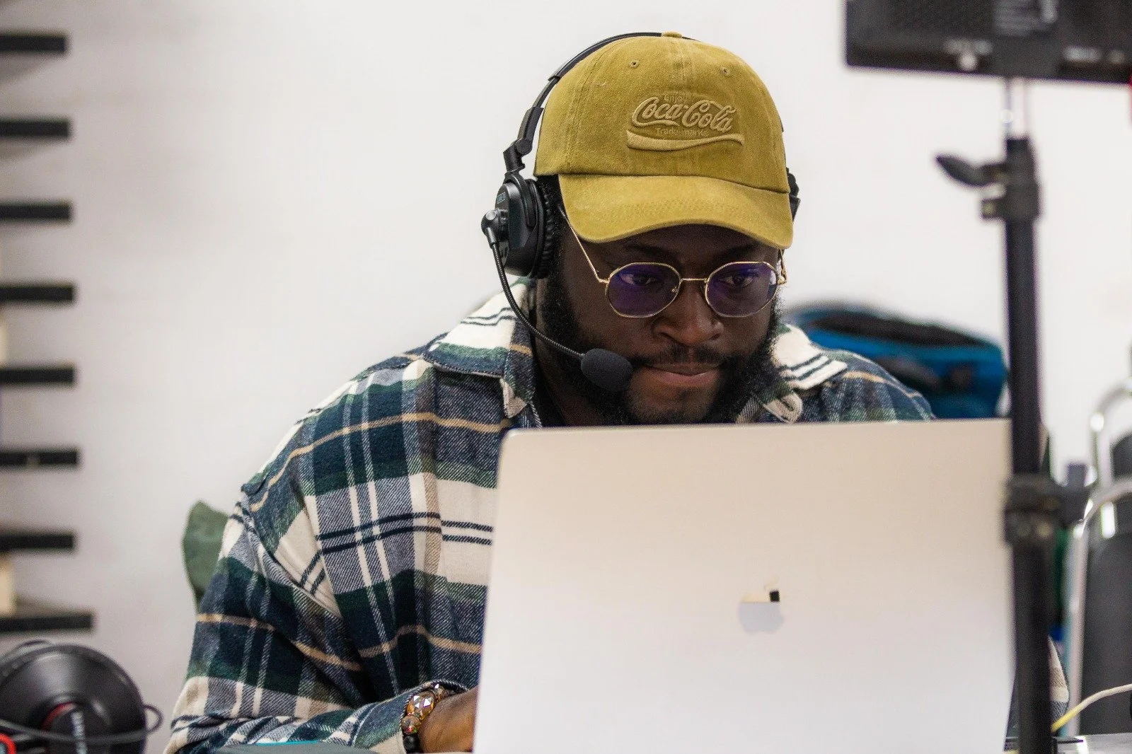 A man wearing a beige Coca-Cola cap, purple-tinted sunglasses, a checkered shirt, and a headset with a microphone is working on a laptop in an office environment.