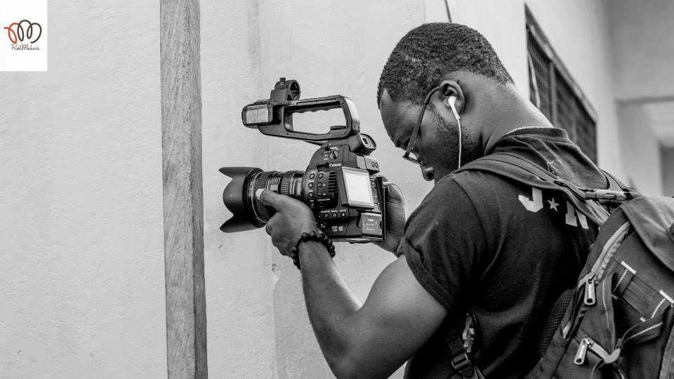 A man taking a photo with a professional camera mounted on a stabilizer, wearing glasses and earphones, carrying a backpack, standing against a wall.