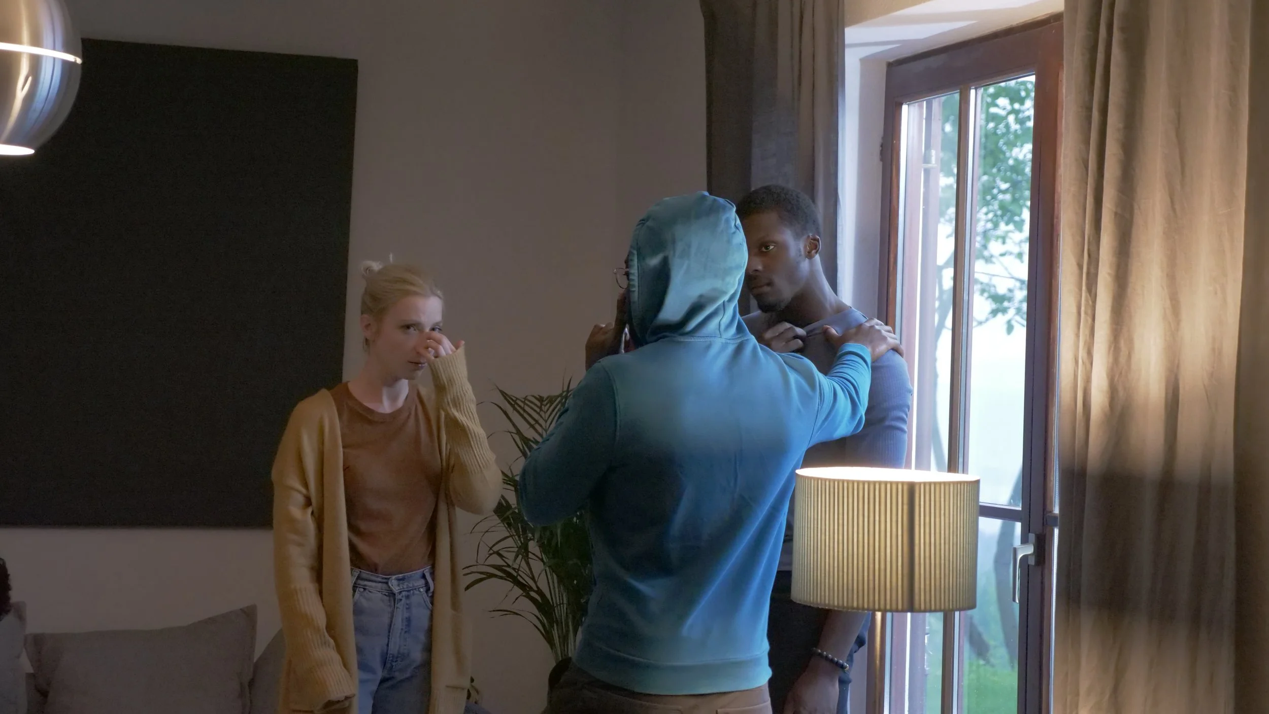 Three people in a room near a window: a woman with blonde hair and a tan sweater, a man with a blue hoodie, and a man with a gray shirt. The woman appears to be wiping her eye, and the man in the gray shirt is looking at the man in the hoodie, who is adjusting his hoodie.