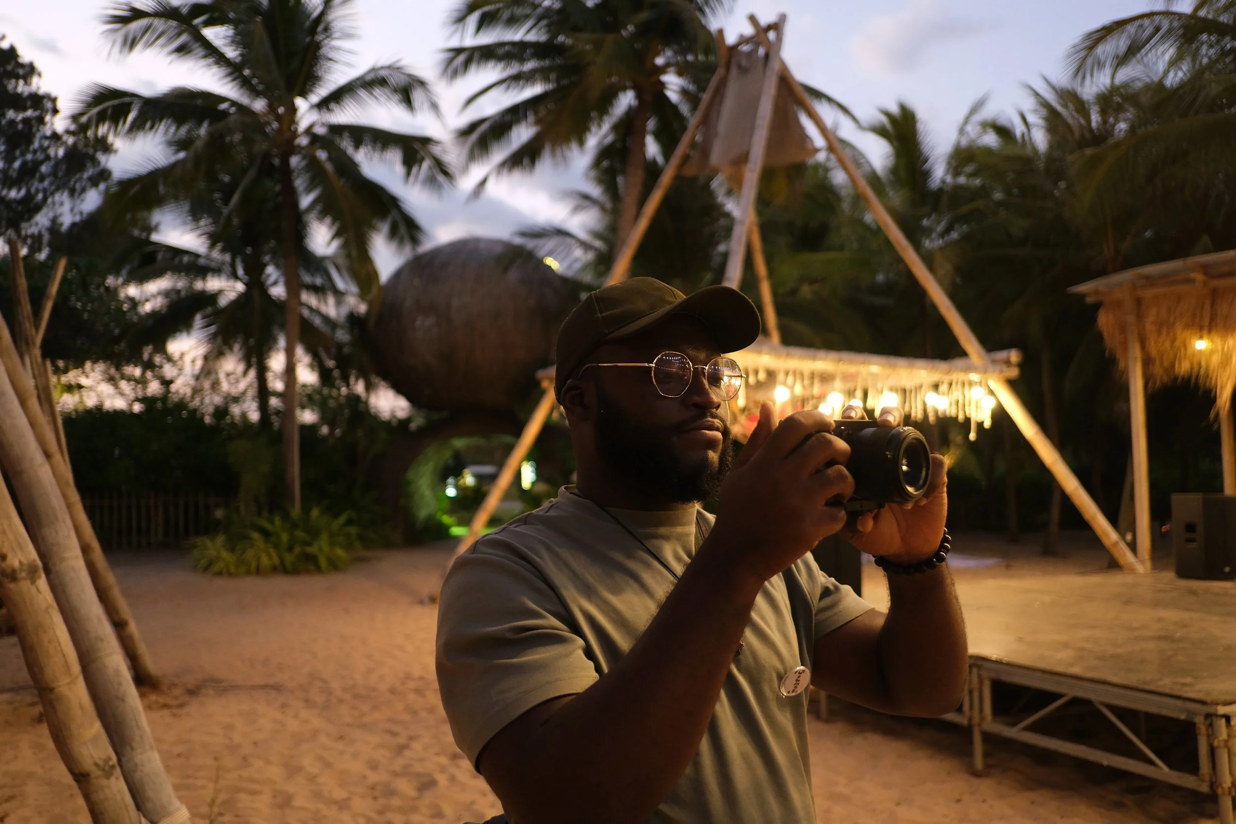 A man with glasses and a cap, holding a camera, standing outdoors at dusk with palm trees and wooden structures in the background.