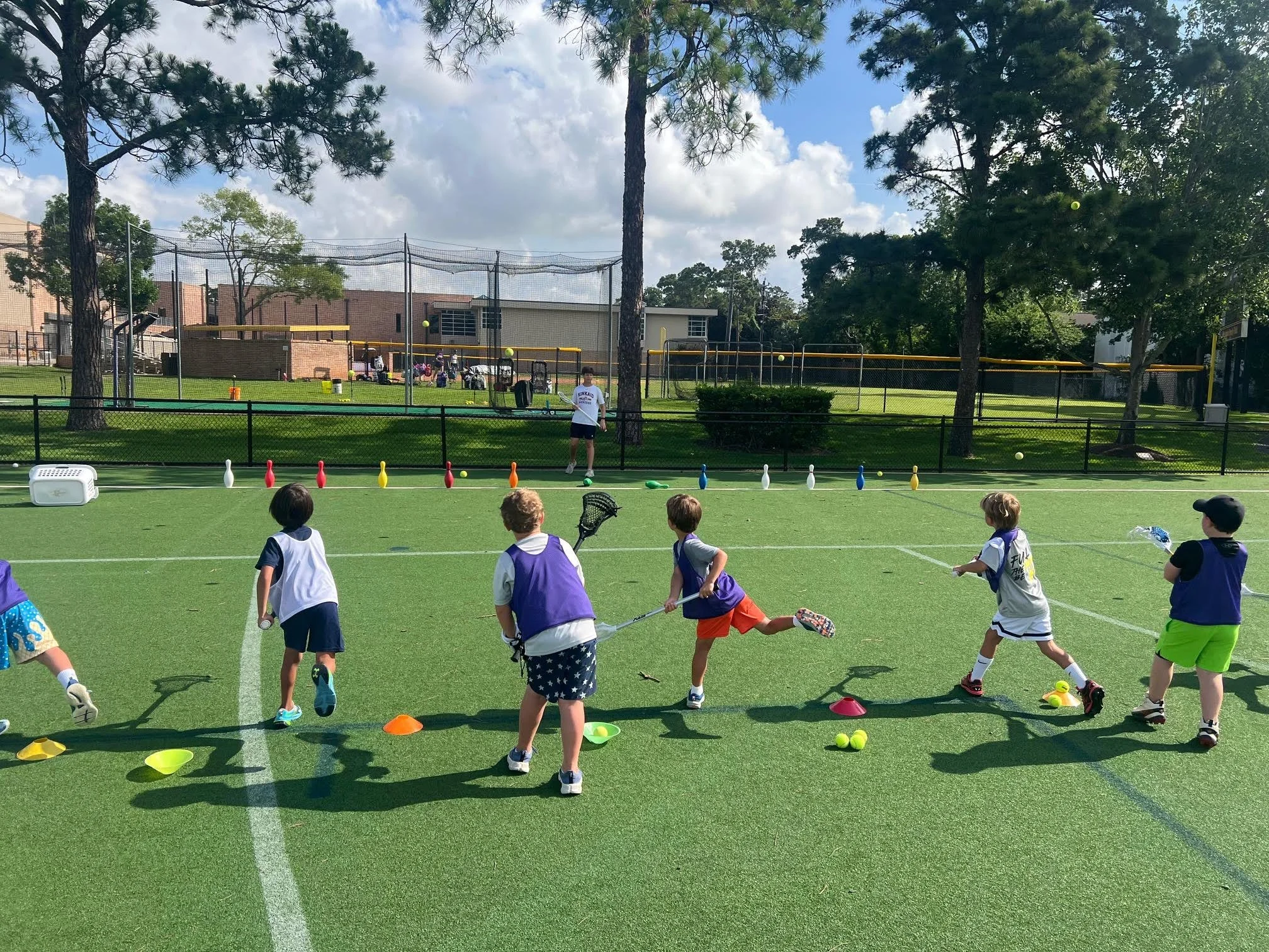 Children participating in a field day event on a green field, with cones, balls, and relay race equipment set up. A coach or teacher stands near a netted tennis court, with trees and a building in the background, under a partly cloudy sky.
