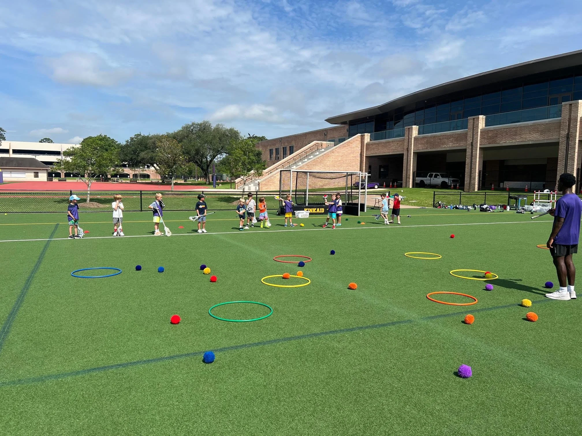 Children and a coach practicing lacrosse on a sports field with colorful hoops and balls.