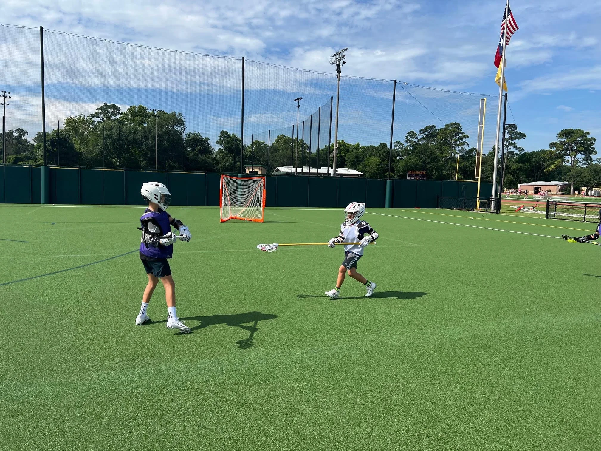 Two young children wearing helmets and playing lacrosse on a green sports field, with a goal and fence in the background, under a partly cloudy sky.