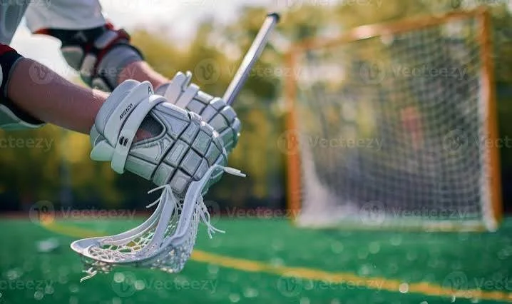 Lacrosse goalie gloves holding a lacrosse stick on a turf field with a lacrosse goal in the background.