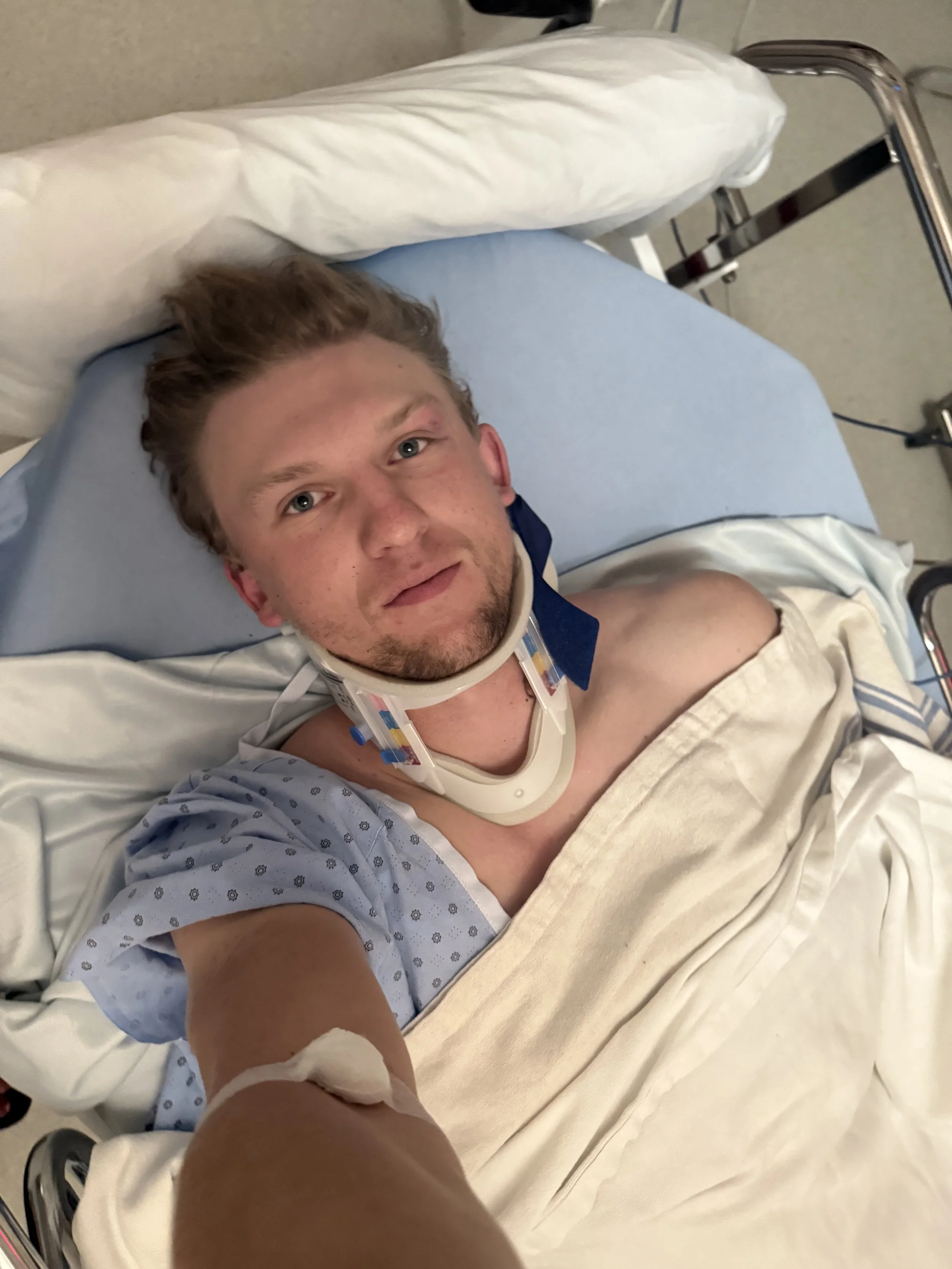 Dylan lying in a hospital bed, wearing a hospital gown, looking up at the camera, with a pillow behind his head, and an IV line connected to his arm after breaking his back