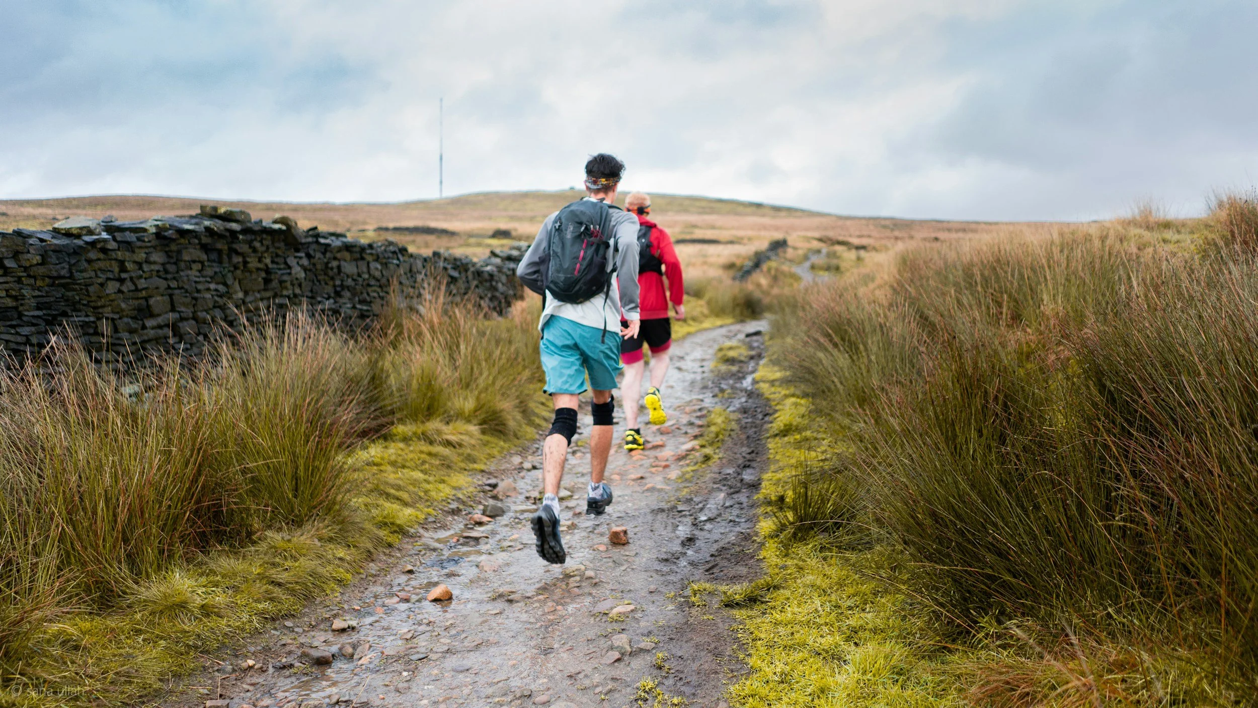 Two people hiking on a muddy trail through grassy landscape under cloudy sky, with stone wall on the side.