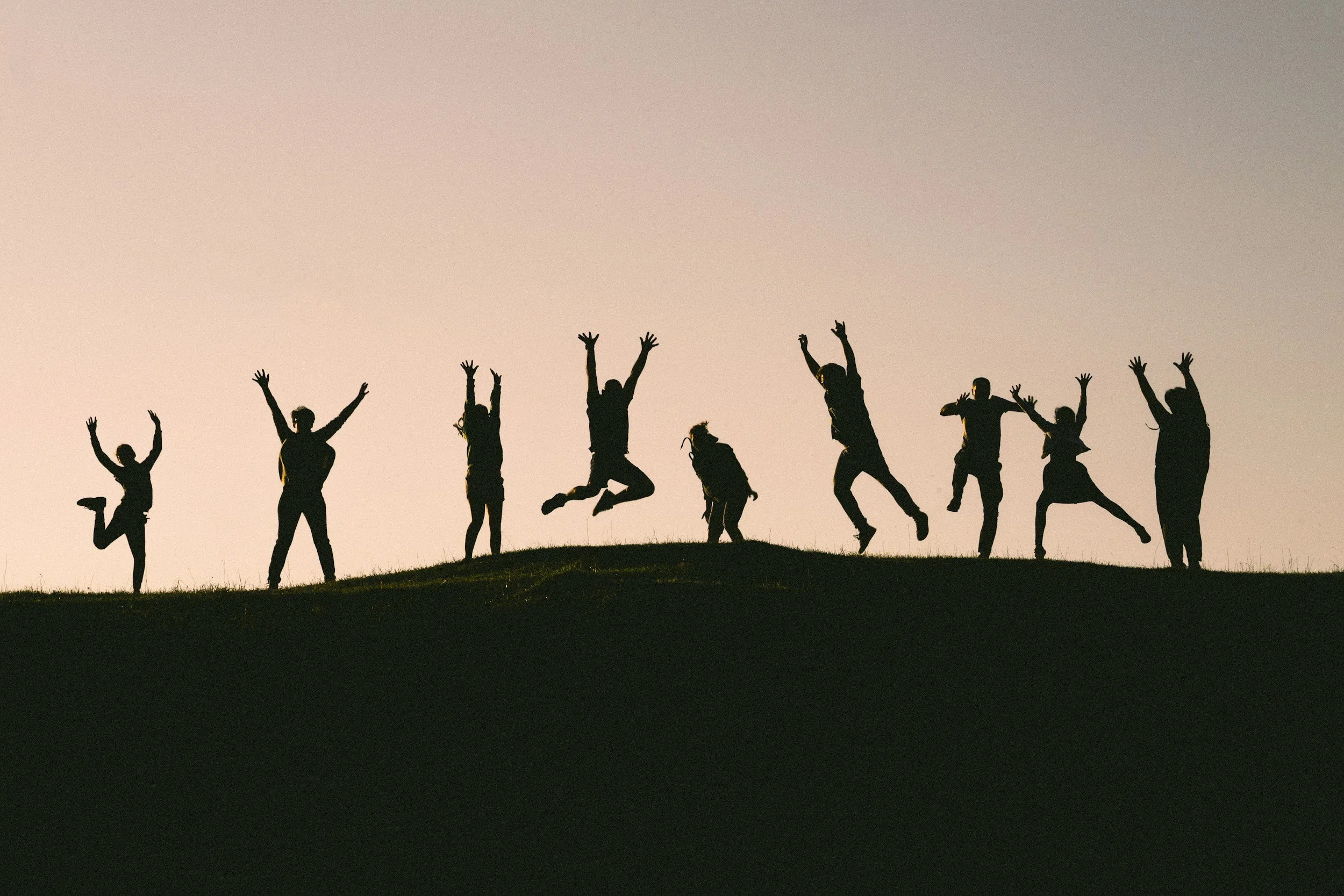 Silhouettes of people jumping and dancing on a grassy hill at sunset.