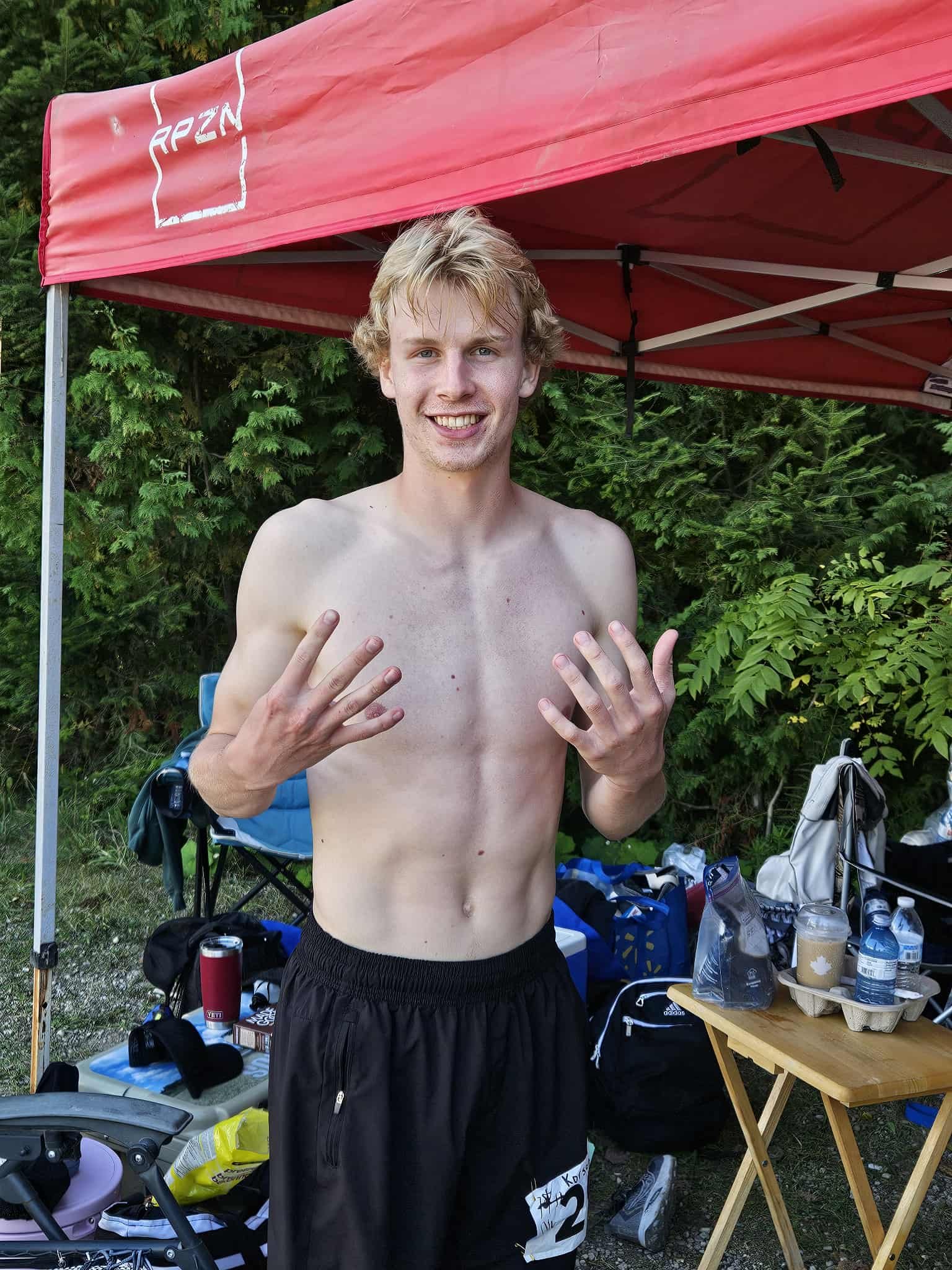 Korson and a smile, shirtless, standing outdoors with nine fingers up representing the 9 laps he finished at his first backyard ultra. Behind him are green foliage and a table with various supplies.
