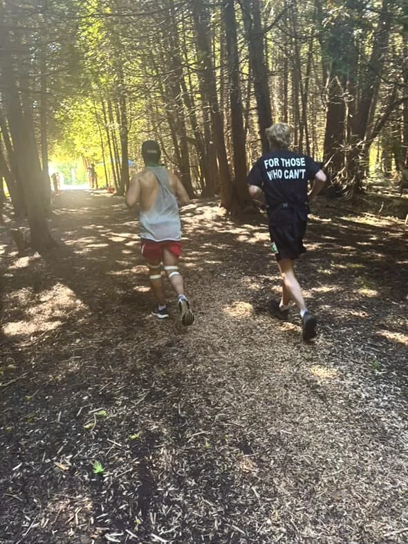Two runners on a wooded trail in the forest, one wearing a gray tank top and red shorts, the other wearing a black shirt with white text and black shorts.
