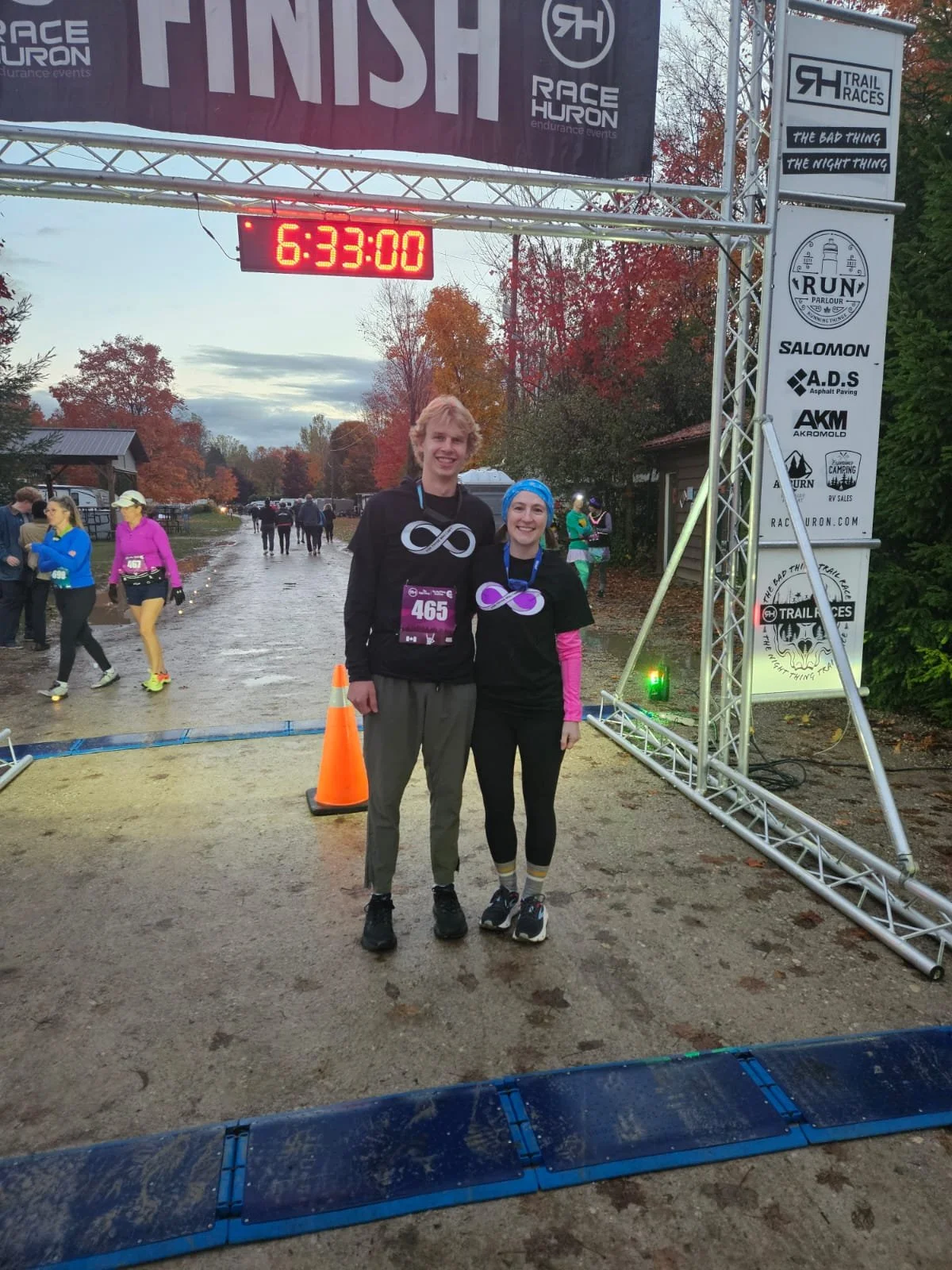 Two smiling runners stand together at the finish line of a race event, with a digital clock showing 6:33:00 in the background and other participants nearby.