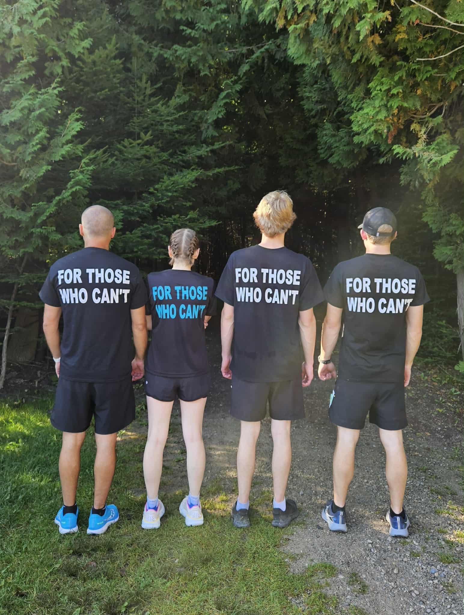Four people standing outdoors with backs facing the camera, wearing black T-shirts that read 'FOR THOSE WHO CAN'T' and black shorts, facing a dense forest of green trees.