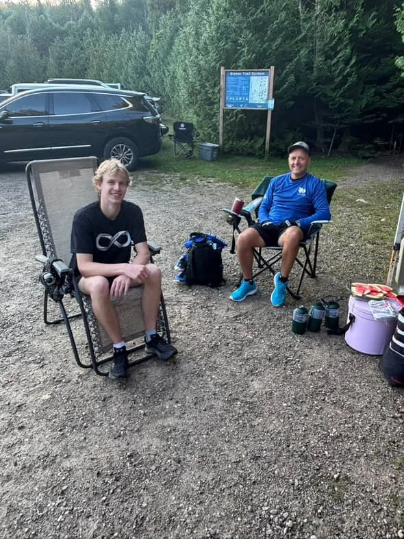 Two people sitting outdoors near parked cars, surrounded by trees, with camping gear and a trail map in the background.