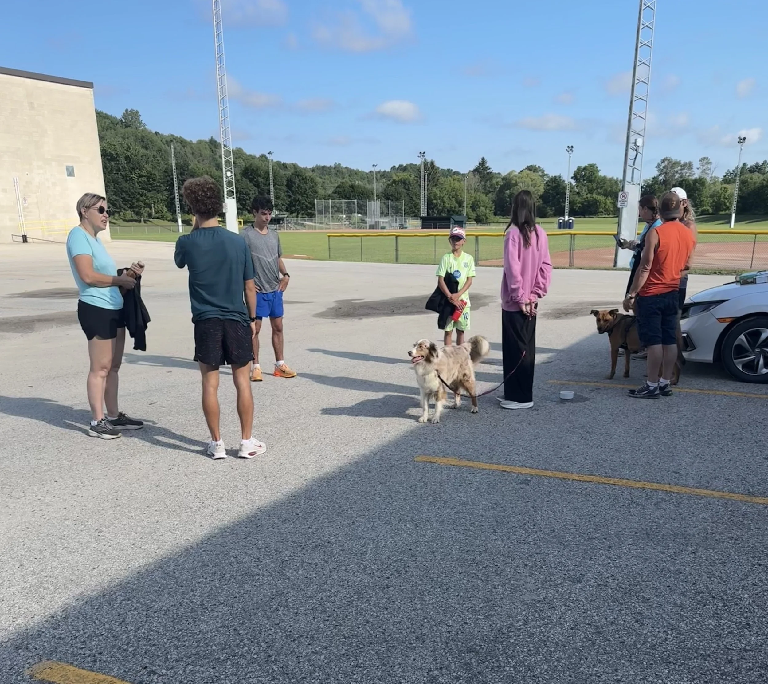 Group of the run-er walk club standing and chatting in a parking lot near a baseball field on a sunny day, with clear blue sky and green trees in the background.