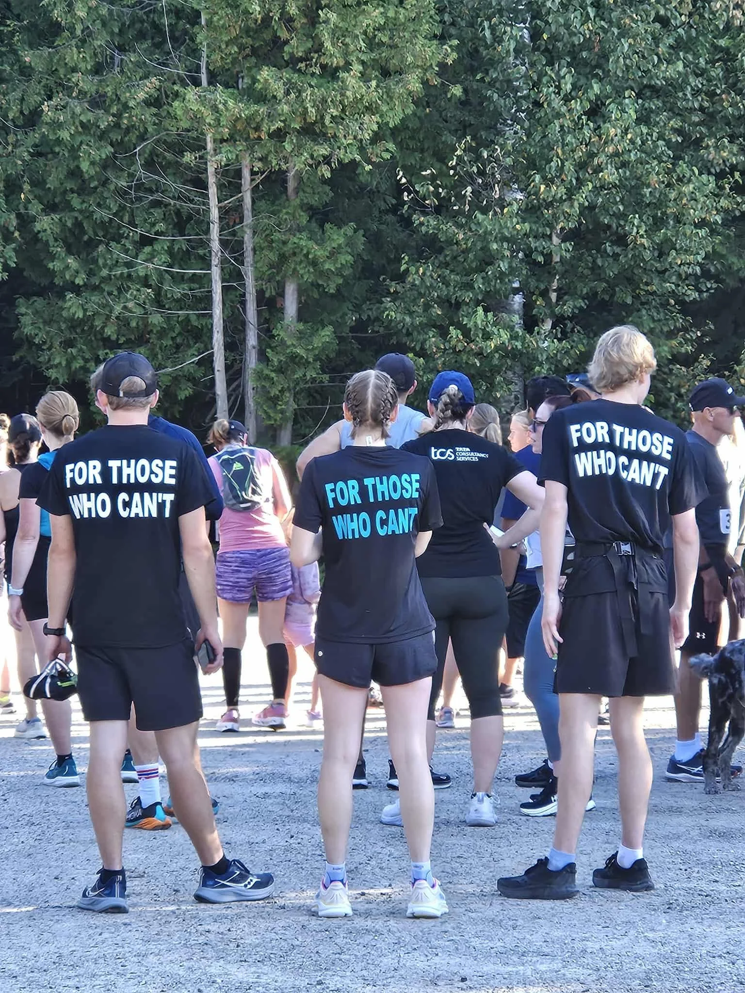 Group of people gathered outdoors on a sunny day, wearing athletic clothing, with some wearing shirts that say 'For Those Who Can't', near a wooded area with tall trees and utility poles.