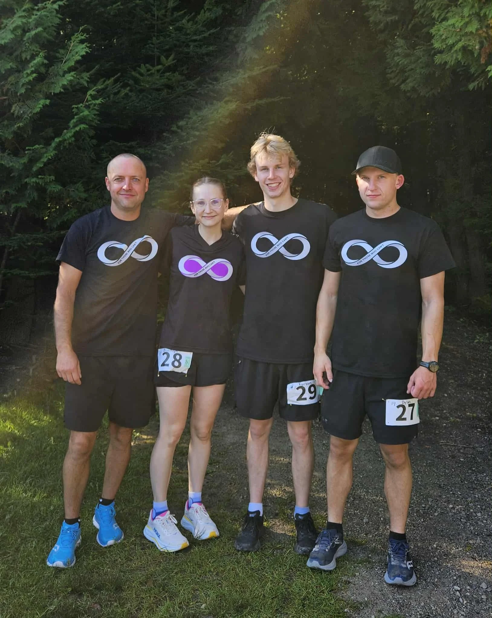 Four people standing outdoors on grass, wearing athletic clothing and running shoes, with bib numbers attached, suggesting they are participating in a race or running event, surrounded by trees.