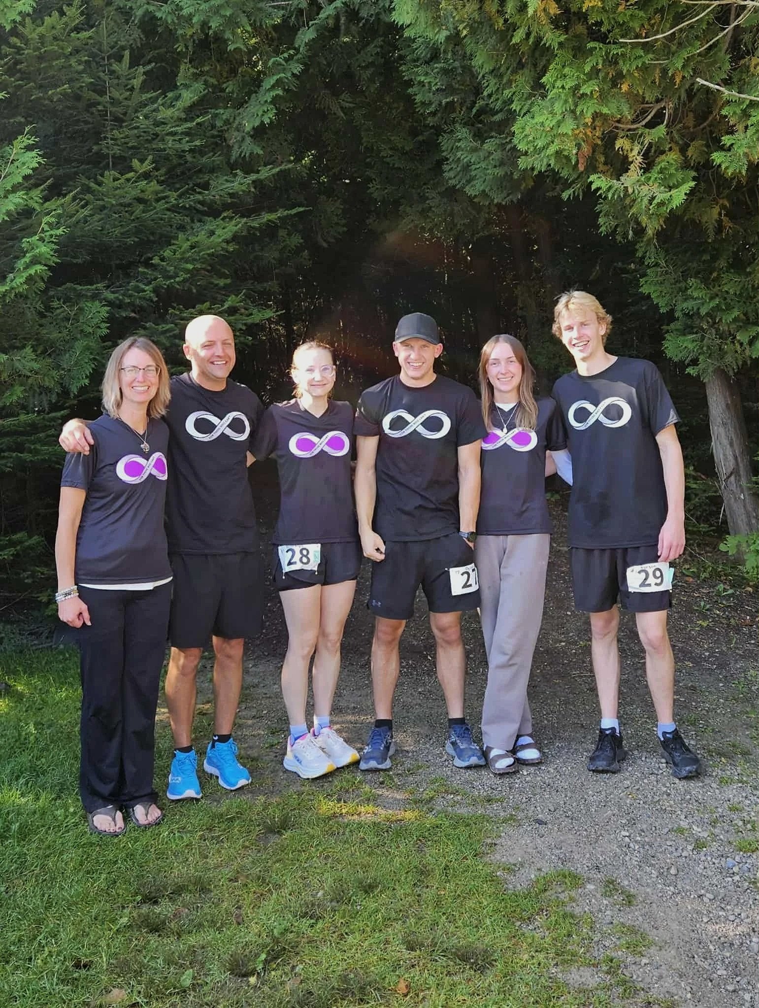Group of six people standing outdoors in front of green trees, smiling, wearing matching black shirts with a white infinity symbol logo, and numbered bibs.