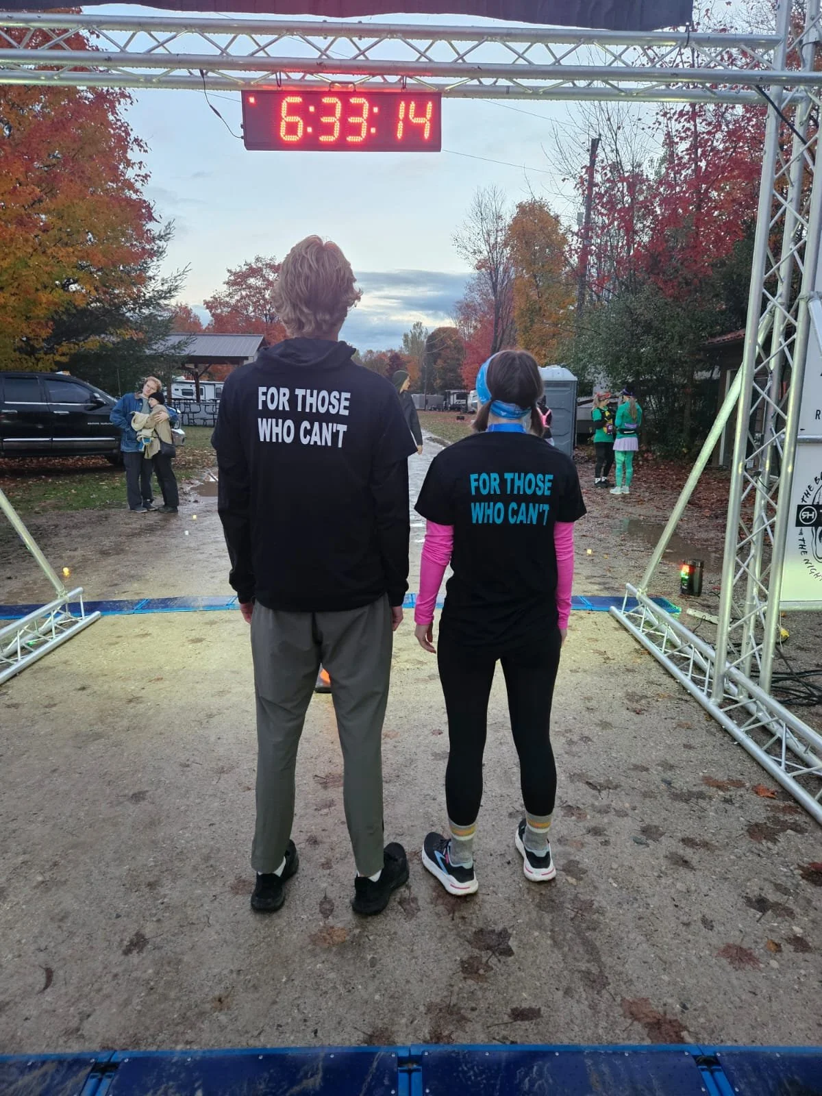 Two people, a man and a woman, wearing matching black shirts with the phrase "FOR THOSE WHO CAN'T" printed on the back, are standing at the start or finish line of a race. There is a digital clock above them displaying the time 6:33:14. The scene app