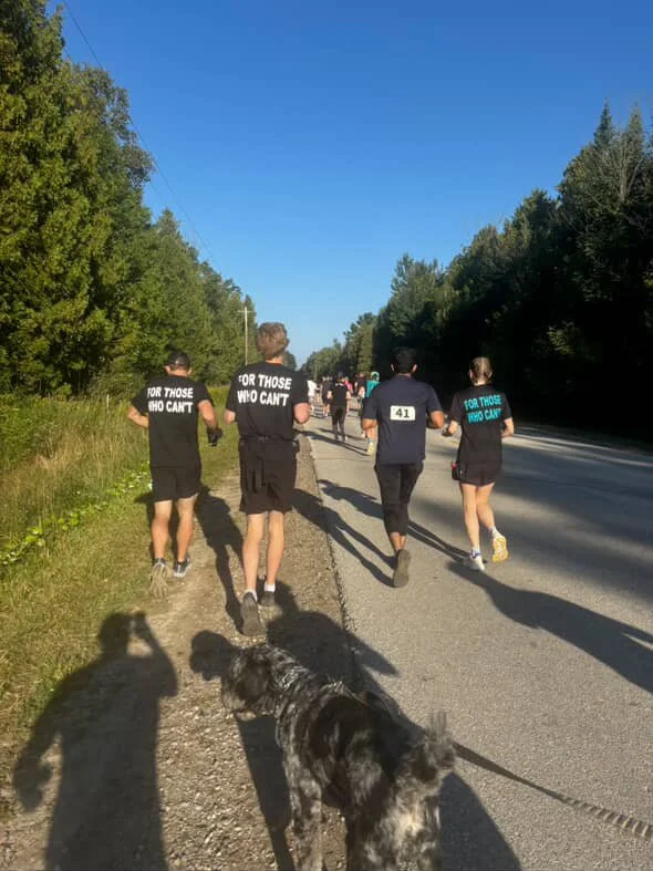People participating in a running event on a road lined with trees, wearing shirts with the text 'For Those Who Can't.'