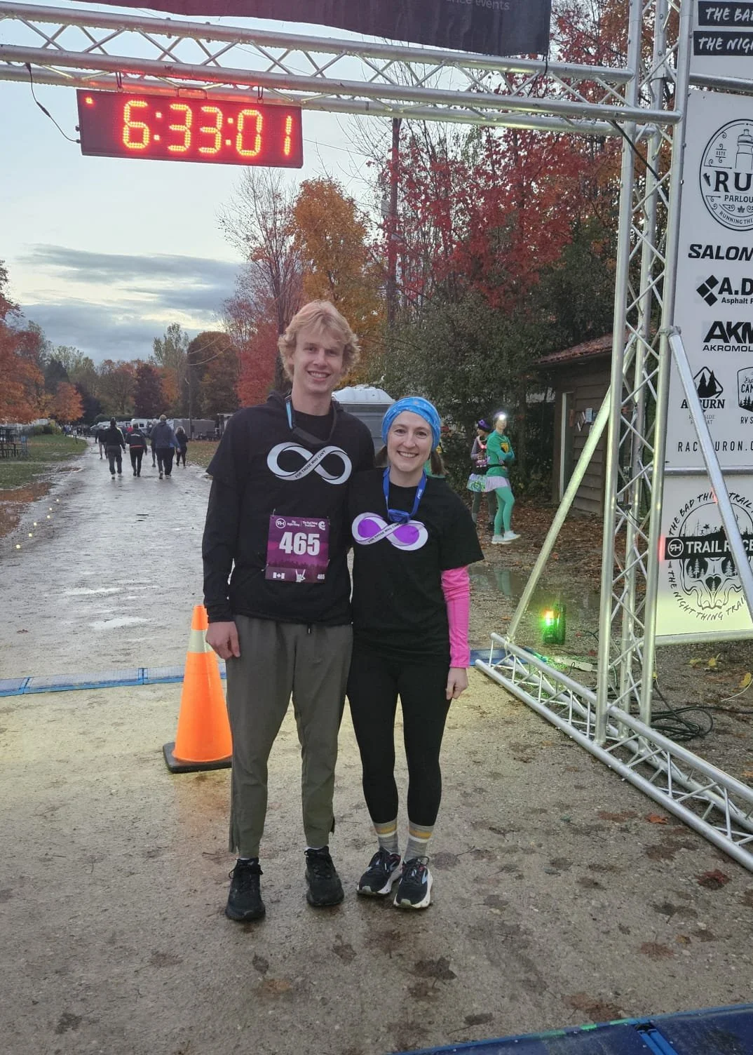 Two cousins smiling, standing at the finish line getting ready and warmed up for a race, under a digital clock showing 6:33:01, surrounded by fall foliage and event banners.