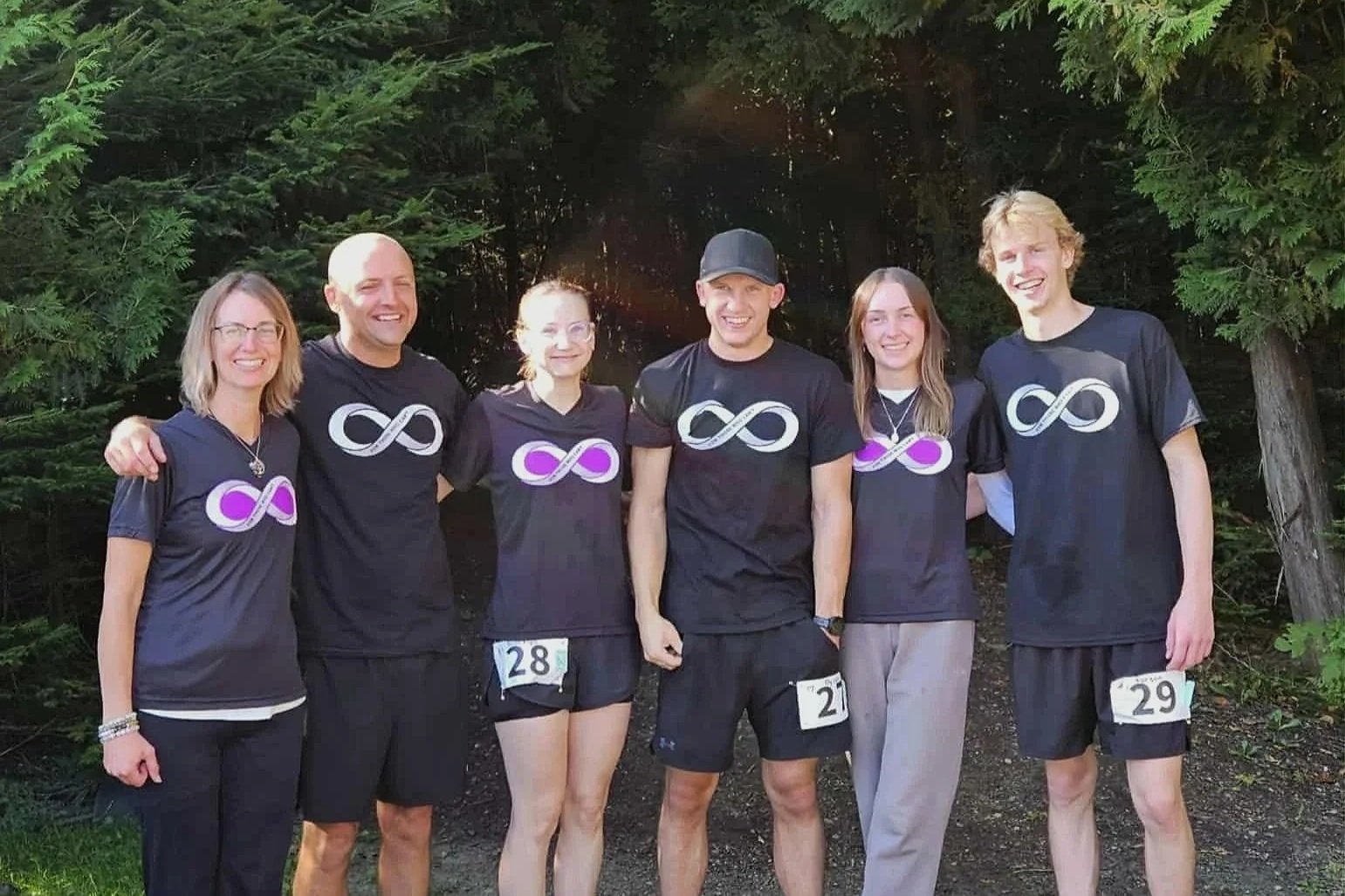 The original six for those who can't supporters, wearing matching black shirts with infinity symbol logos. They are smiling and have race bibs pinned to their shorts or pants. The background features green trees and bushes.