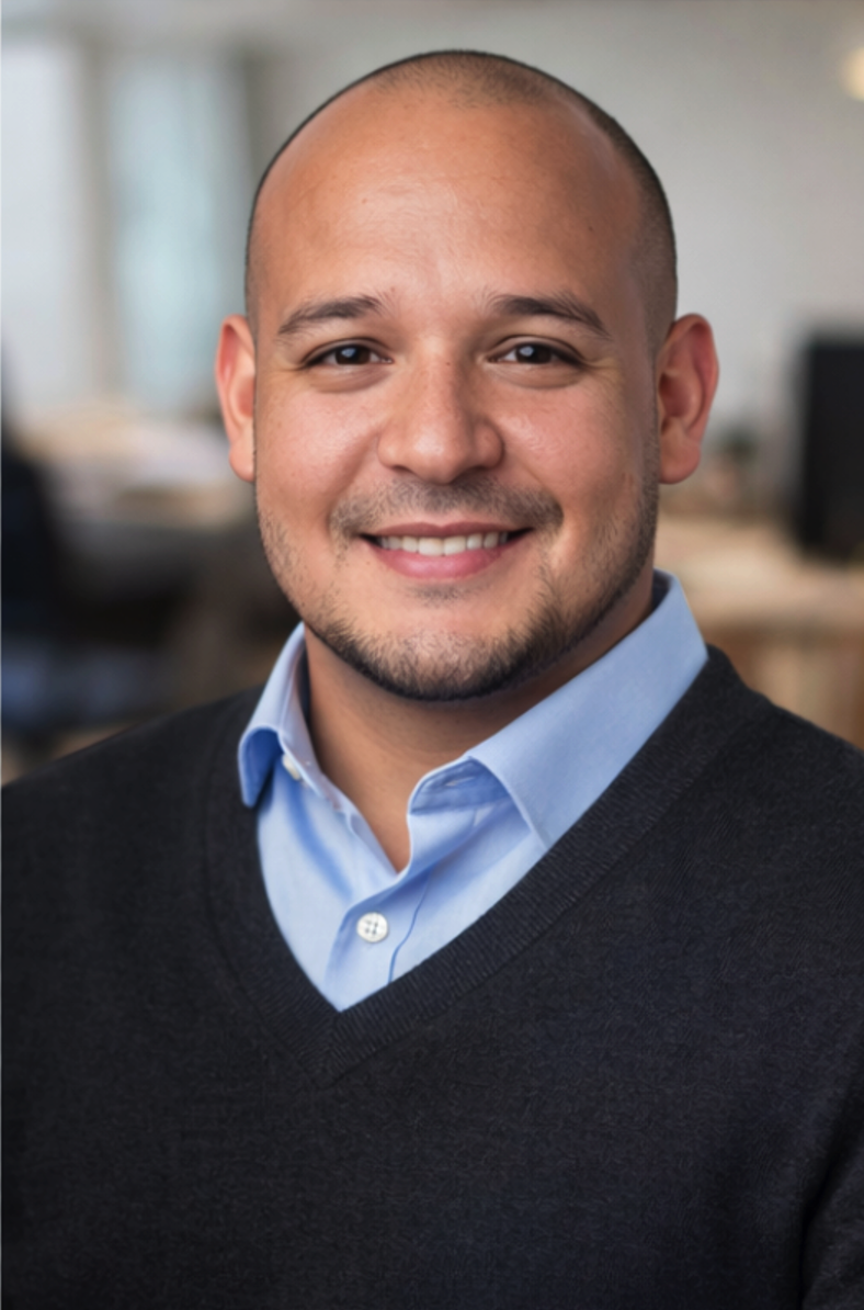 A smiling man with a shaved head wearing a blue collared shirt and a black sweater, in an office setting.