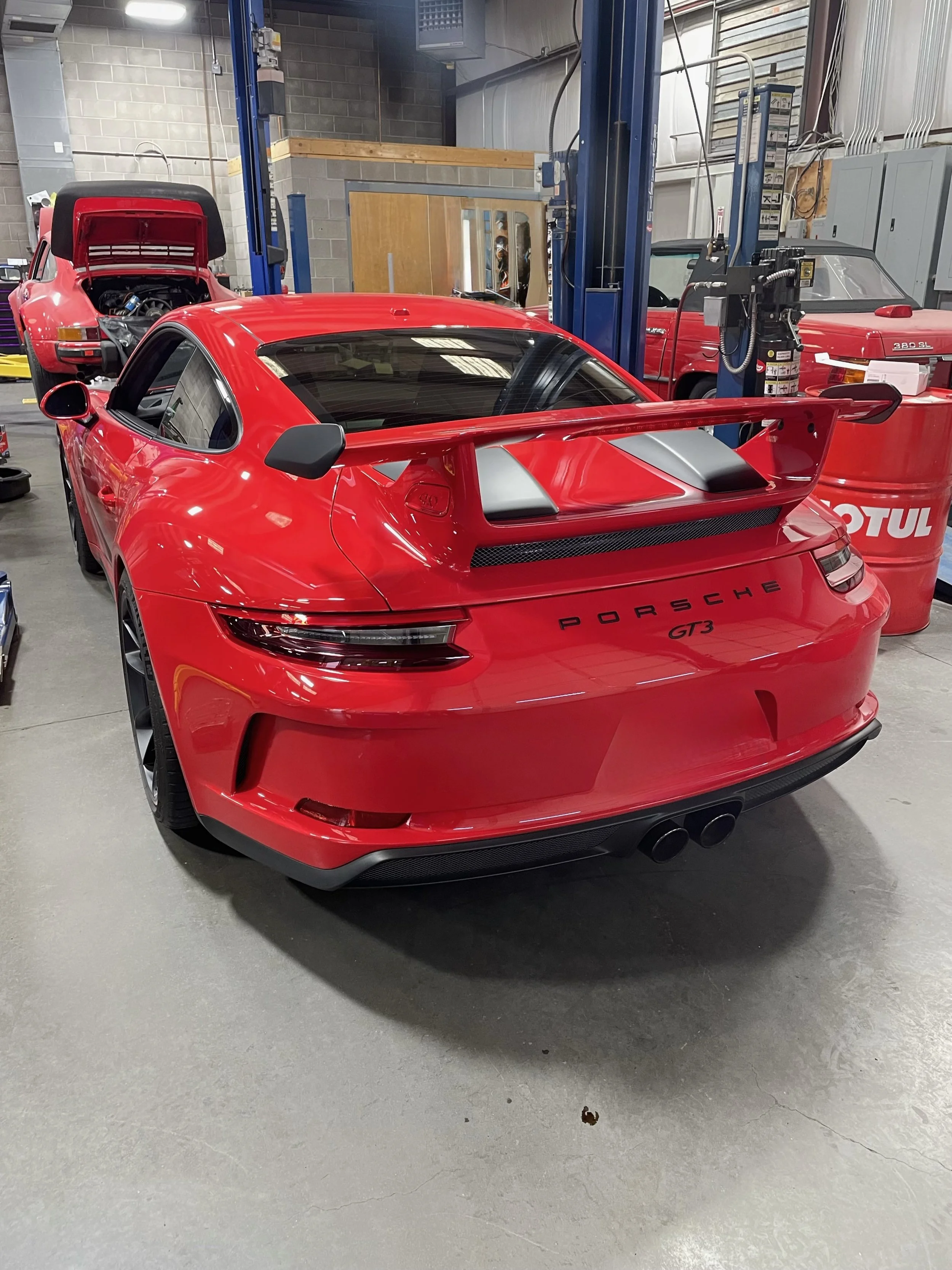 Red Porsche GT3 sports car in a garage with a rear wing spoiler, black side mirrors, and dual exhaust pipes, alongside other classic cars and garage equipment.