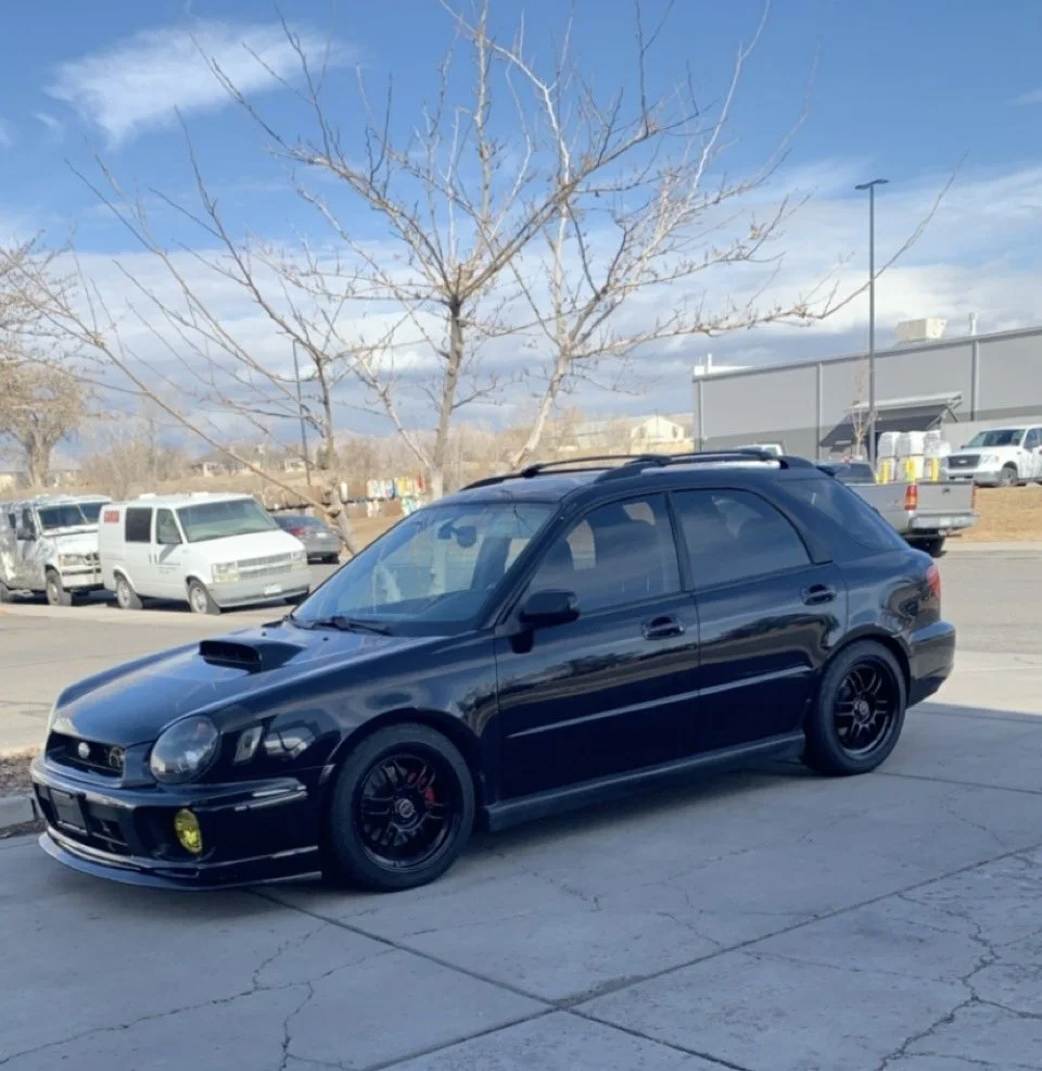 A black Subaru wagon parked on a concrete lot with a tree and other vehicles in the background on a partly cloudy day.