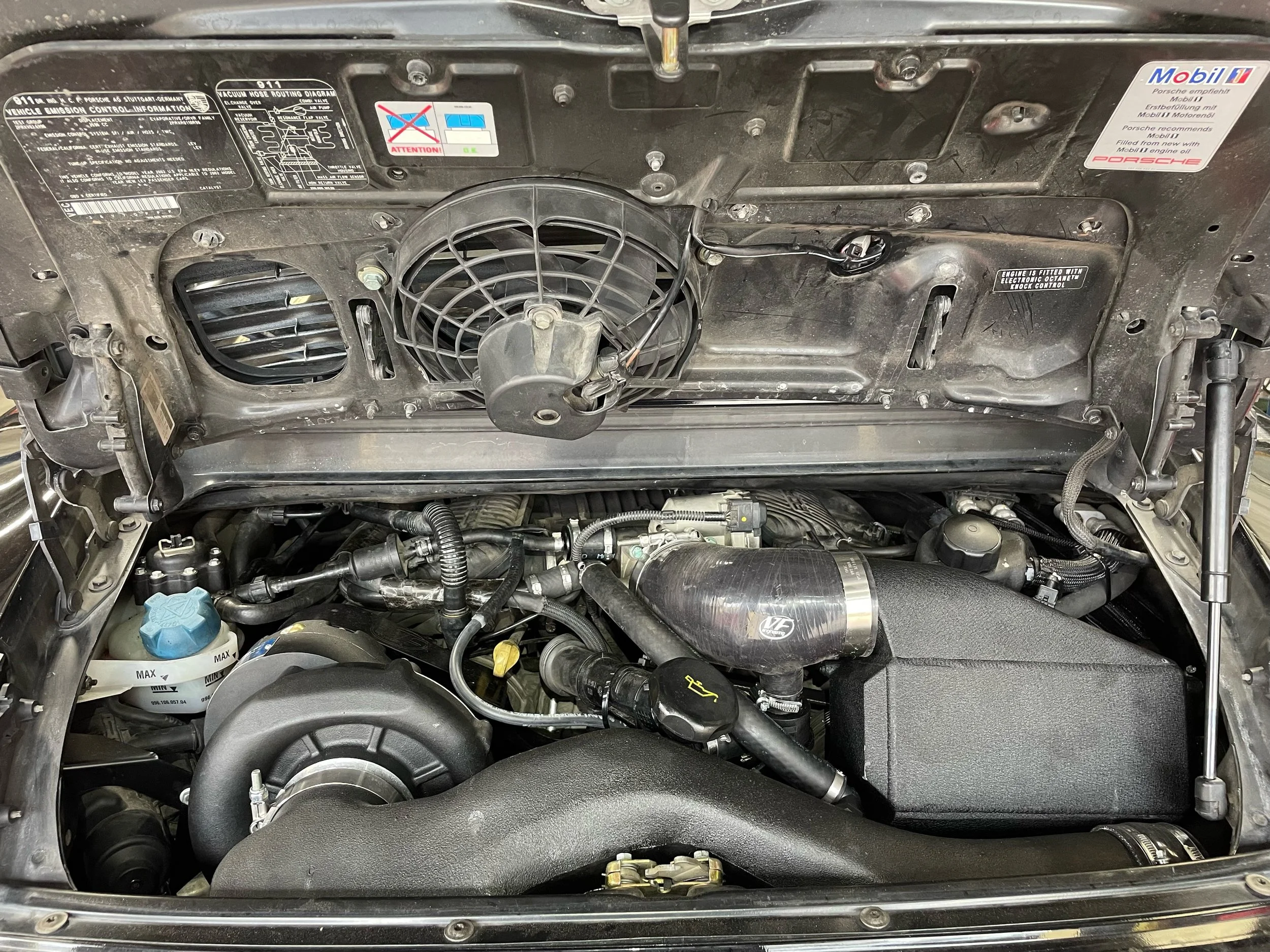 Under the hood of a car showing engine components, including fans, hoses, caps, and an aftermarket Procharger