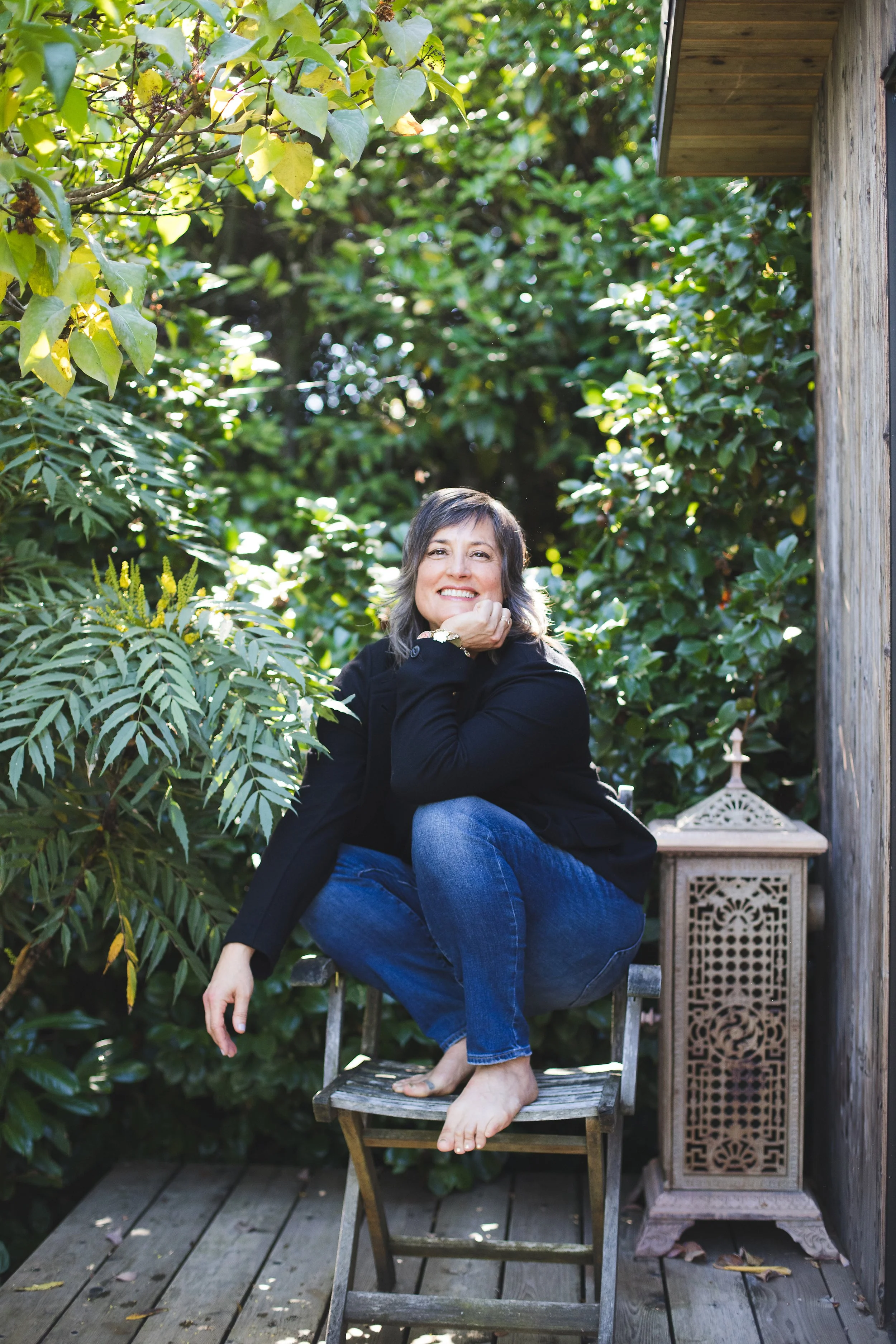 Pema smiling and sitting on a wooden chair outdoors among lush greenery, with a wooden deck and a decorative side table nearby.