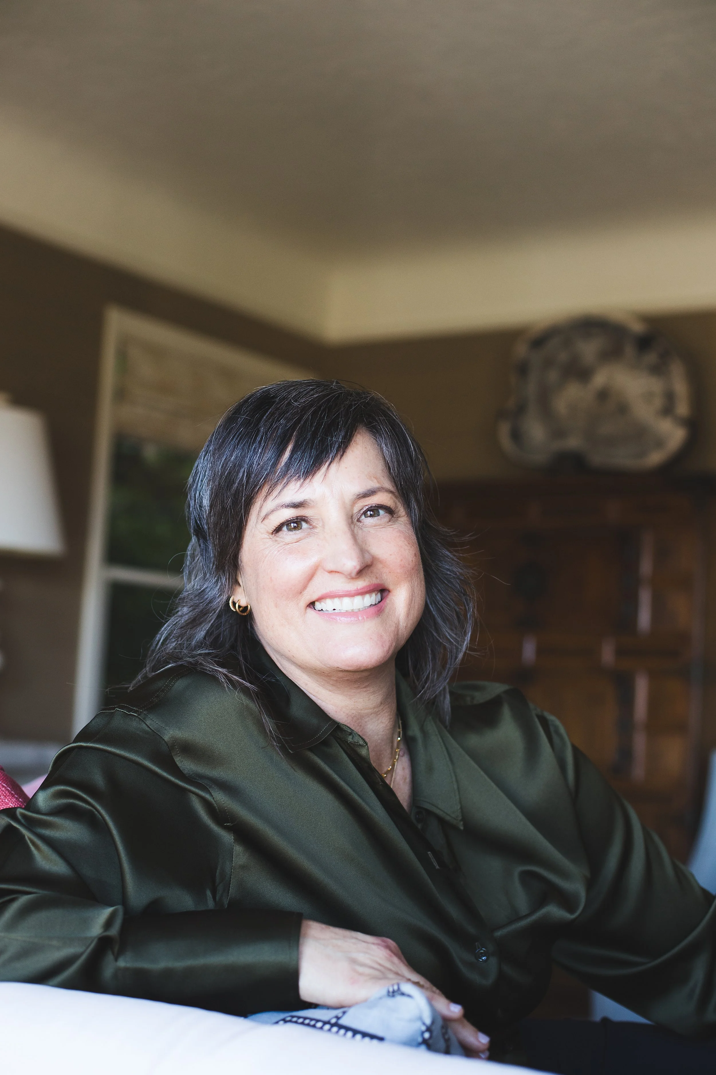 Pema smiling, seated indoors, wearing a dark green, shiny button-up shirt, with a blurred background of a room with art and furniture.