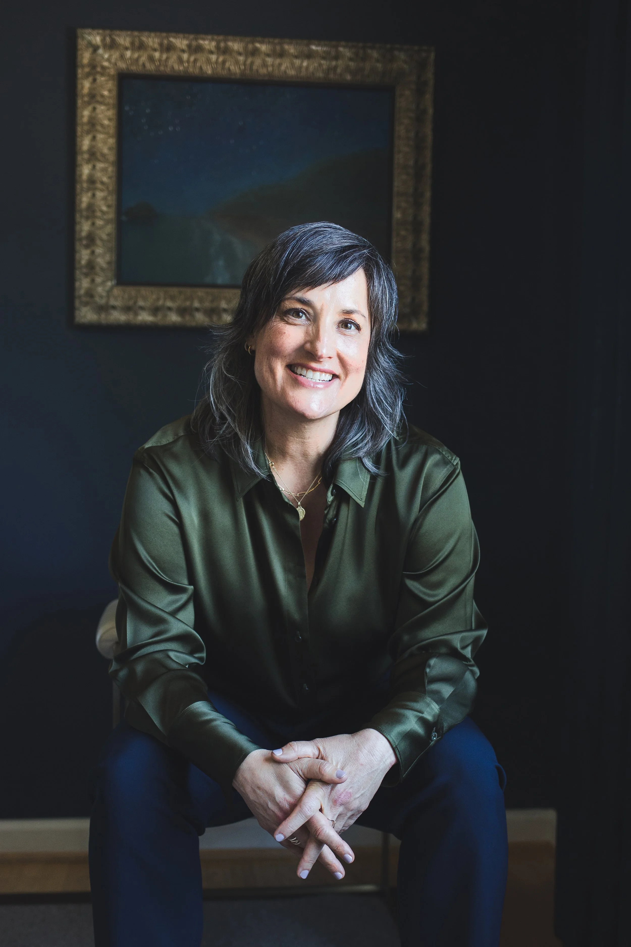 Pema wearing a dark green satin shirt and navy pants sitting on a chair in front of a dark wall with a framed painting of a starry night sky and hills.