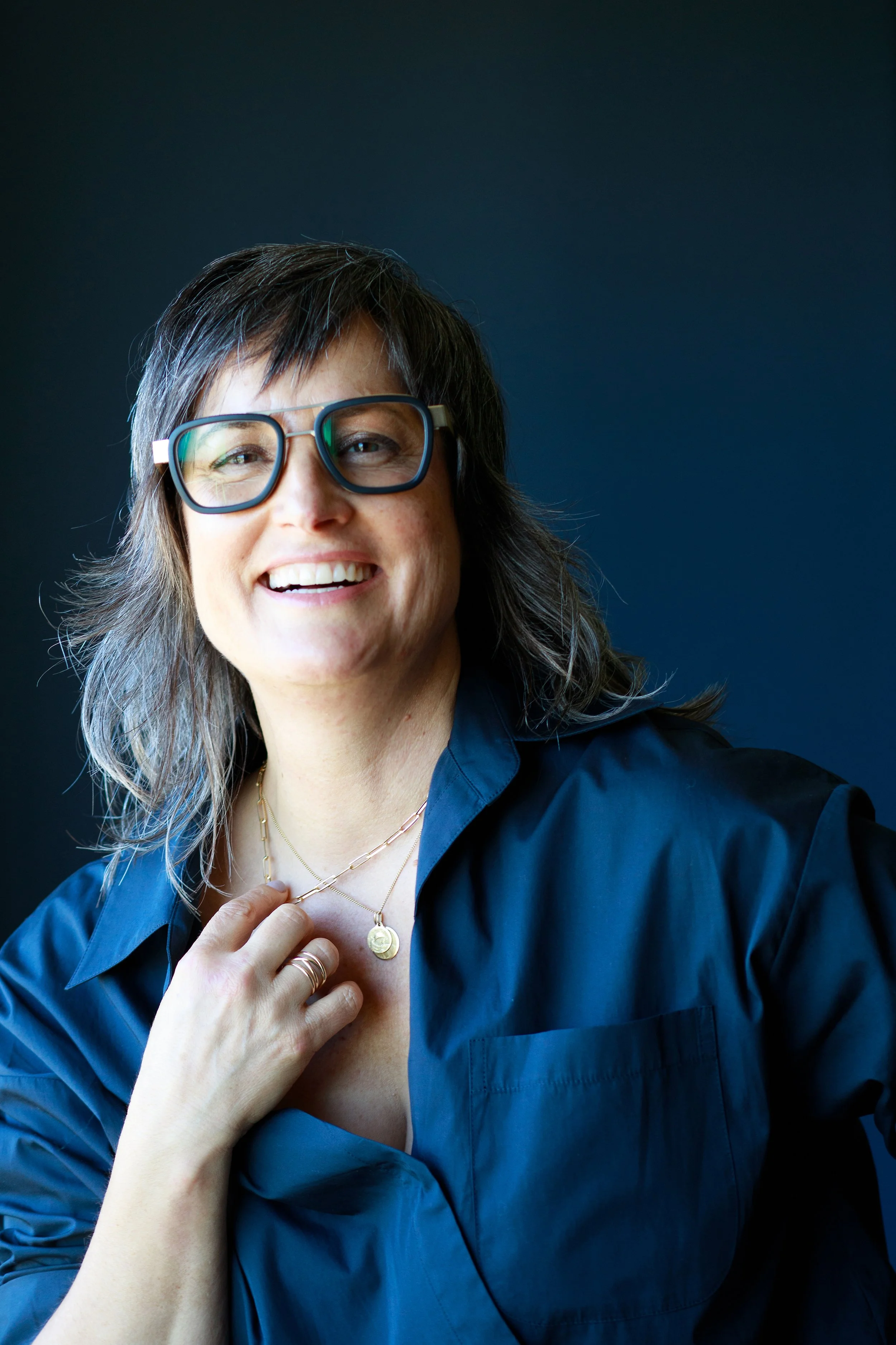 Pema wearing a dark blue shirt, smiling, adjusting a necklace with her hand against a dark background.